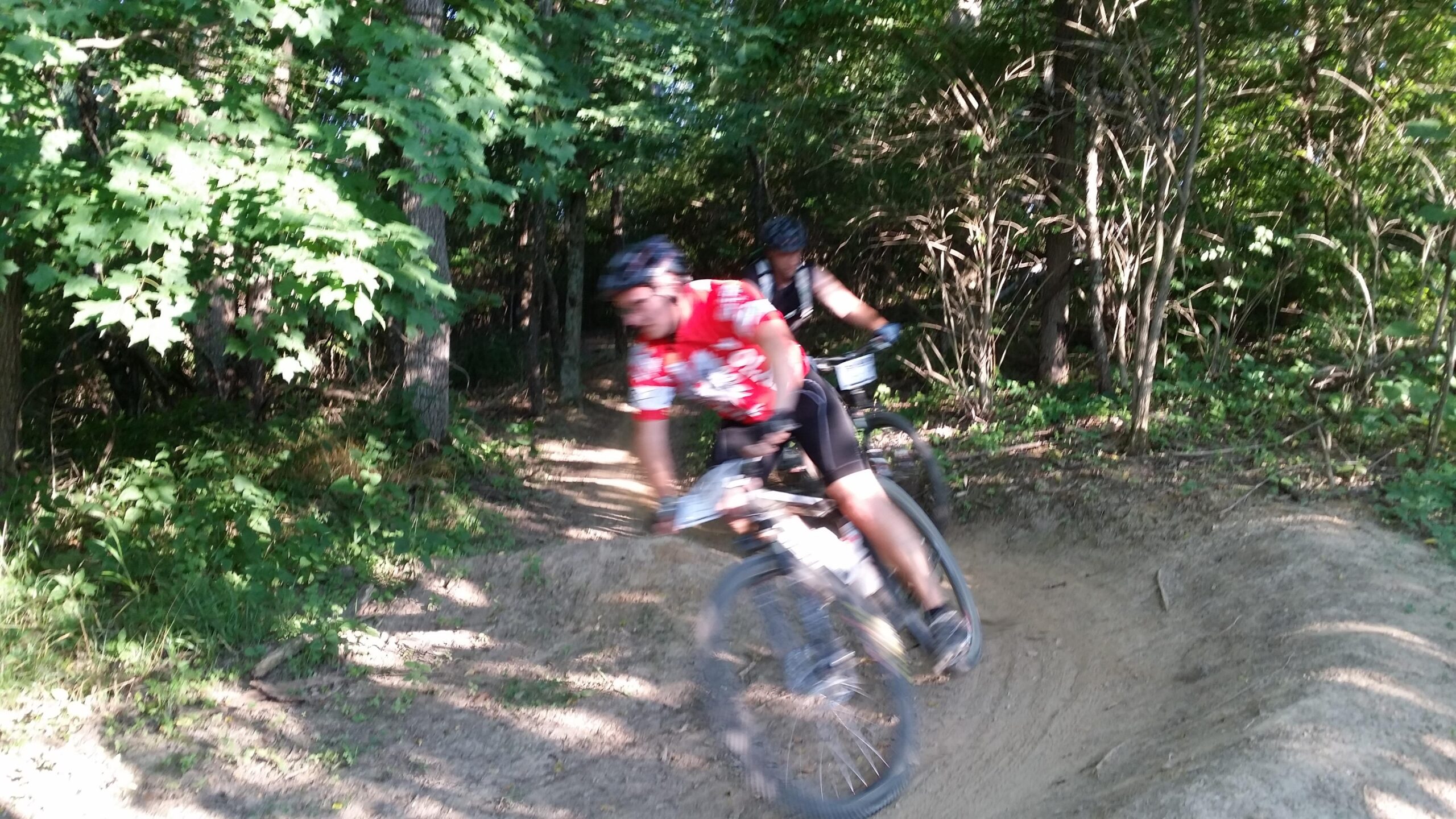 Two mountain bikers navigating a dirt trail through a forested area, with one rider in a red jersey leaning into a turn while the other follows behind. The scene is surrounded by lush green foliage and sunlight filtering through the trees. England Idlewild Mountain Biking Park mountain bike trail.