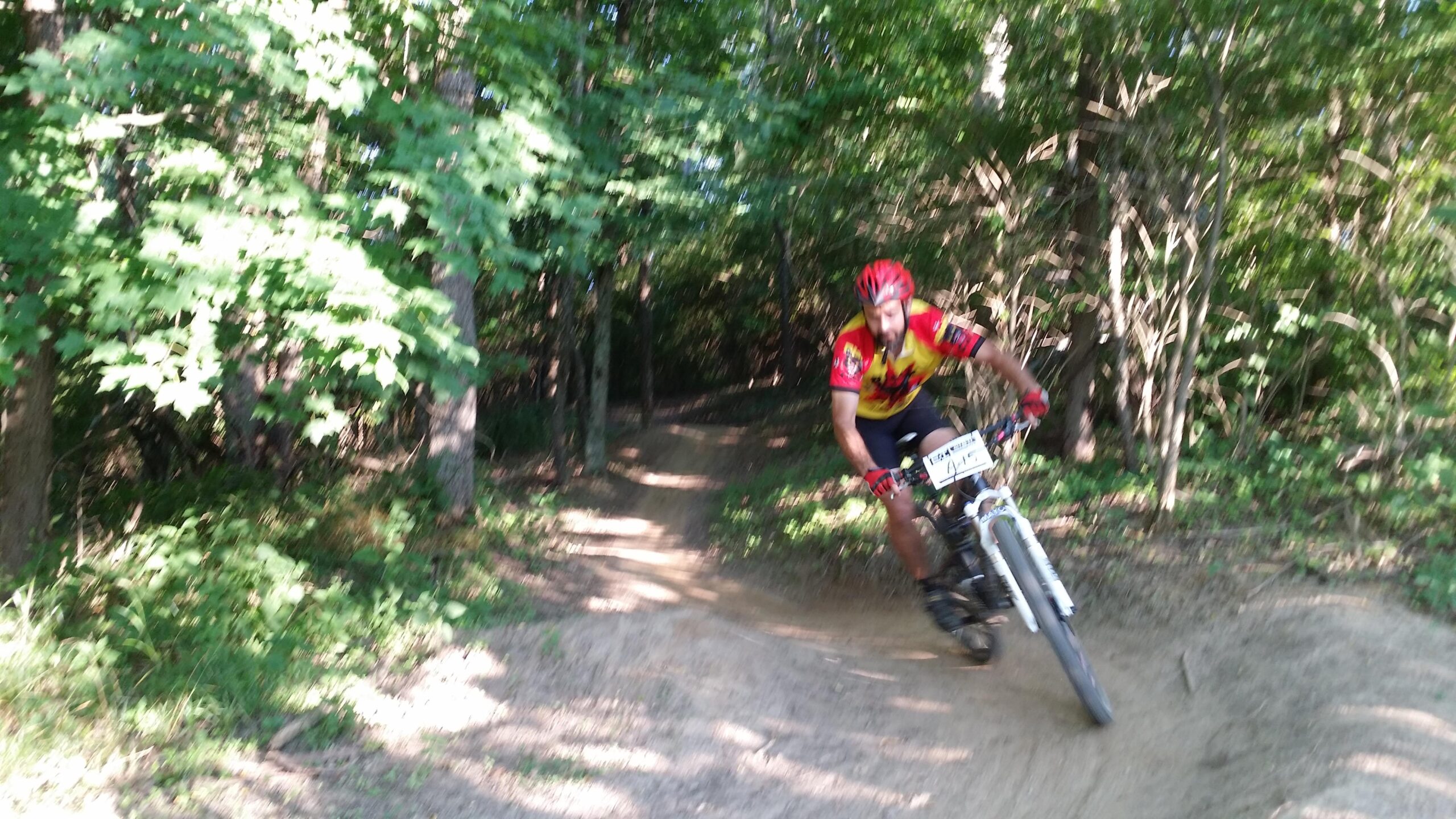 A mountain biker in a red and yellow jersey navigates a dirt trail surrounded by lush green trees, demonstrating agility as he leans into a turn. England Idlewild Mountain Biking Park mountain bike trail.
