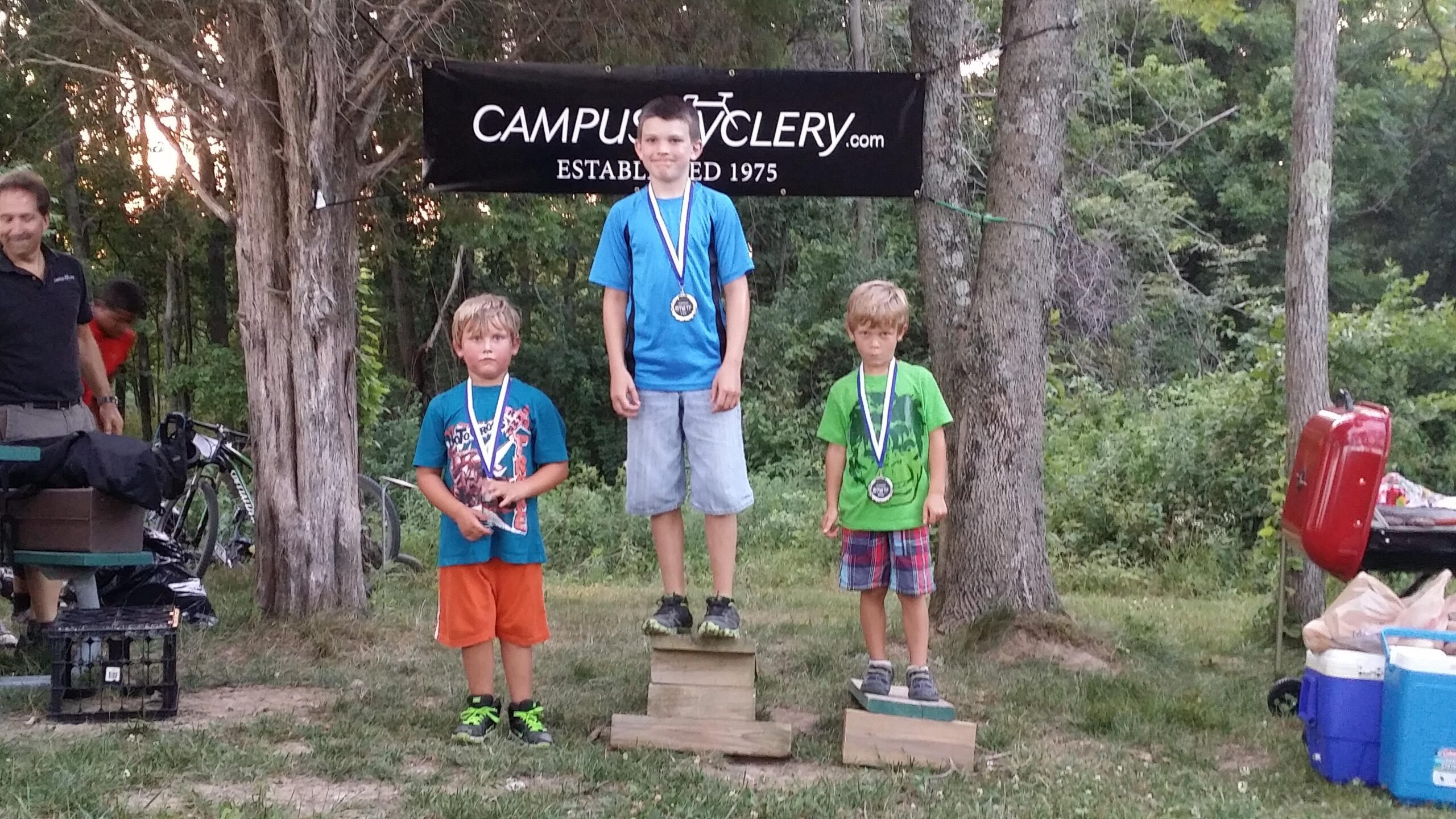 Three young boys stand on a podium, each wearing a medal and displaying varying expressions. The boy in the middle stands on the highest podium step, while the other two are on lower steps. The background features trees and a black banner with the text "CAMPUS CYCLE" and "ESTABLISHED 1975." In the background, an adult is partially visible, and there are bicycles and a grill nearby. The setting appears to be an outdoor event or competition. England Idlewild Mountain Biking Park mountain bike trail.