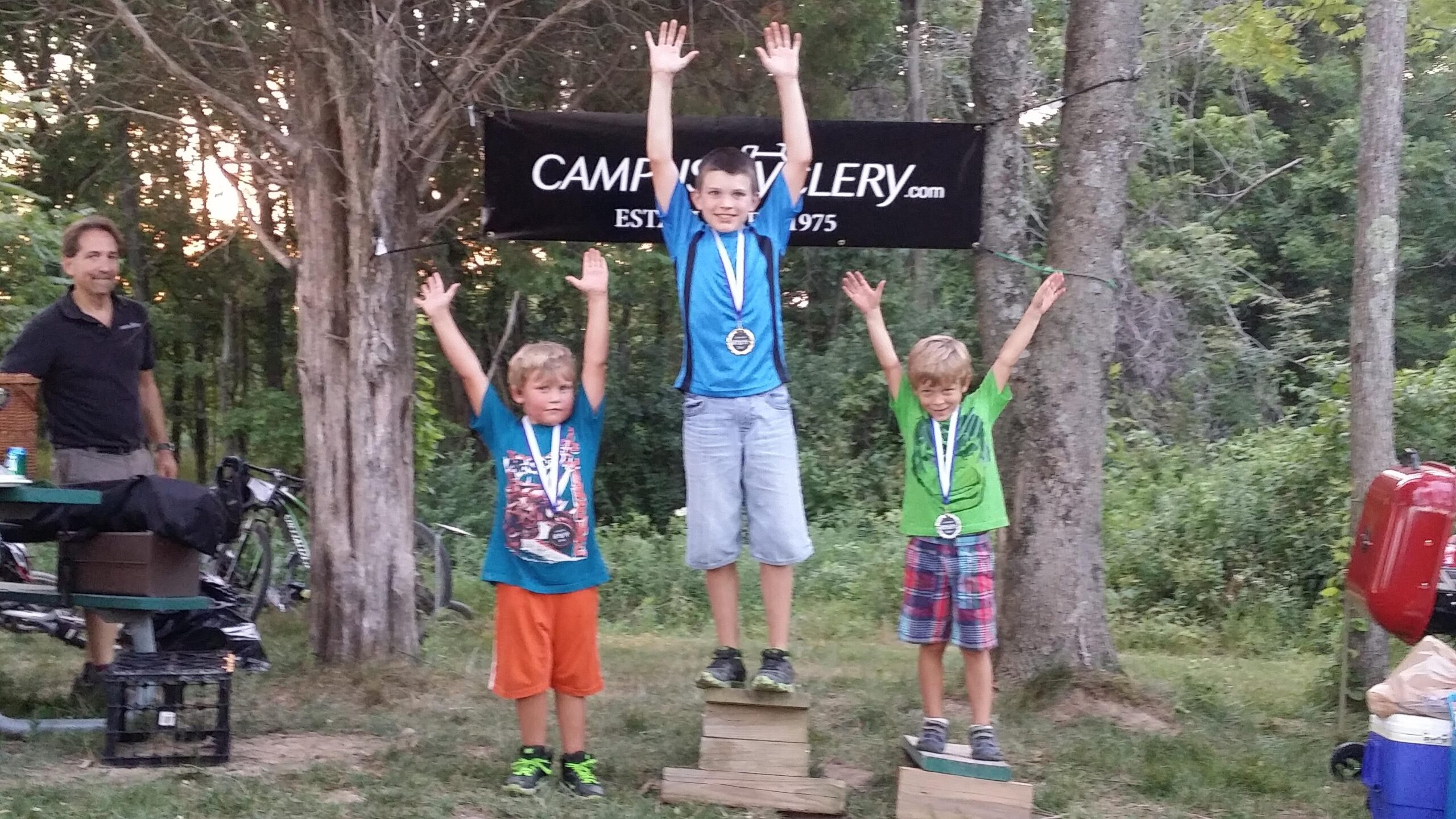 Three children stand on podiums celebrating their achievements, with arms raised in excitement. Each child is wearing a medal around their neck. The boy in the center, wearing a blue shirt, has a medal on a blue ribbon and appears happiest. The child on the left is wearing a blue and orange outfit, and the one on the right is in a green shirt and plaid shorts. In the background, there’s a man standing beside a picnic table, and the scene is set outdoors with trees and a banner in the background. England Idlewild Mountain Biking Park mountain bike trail.