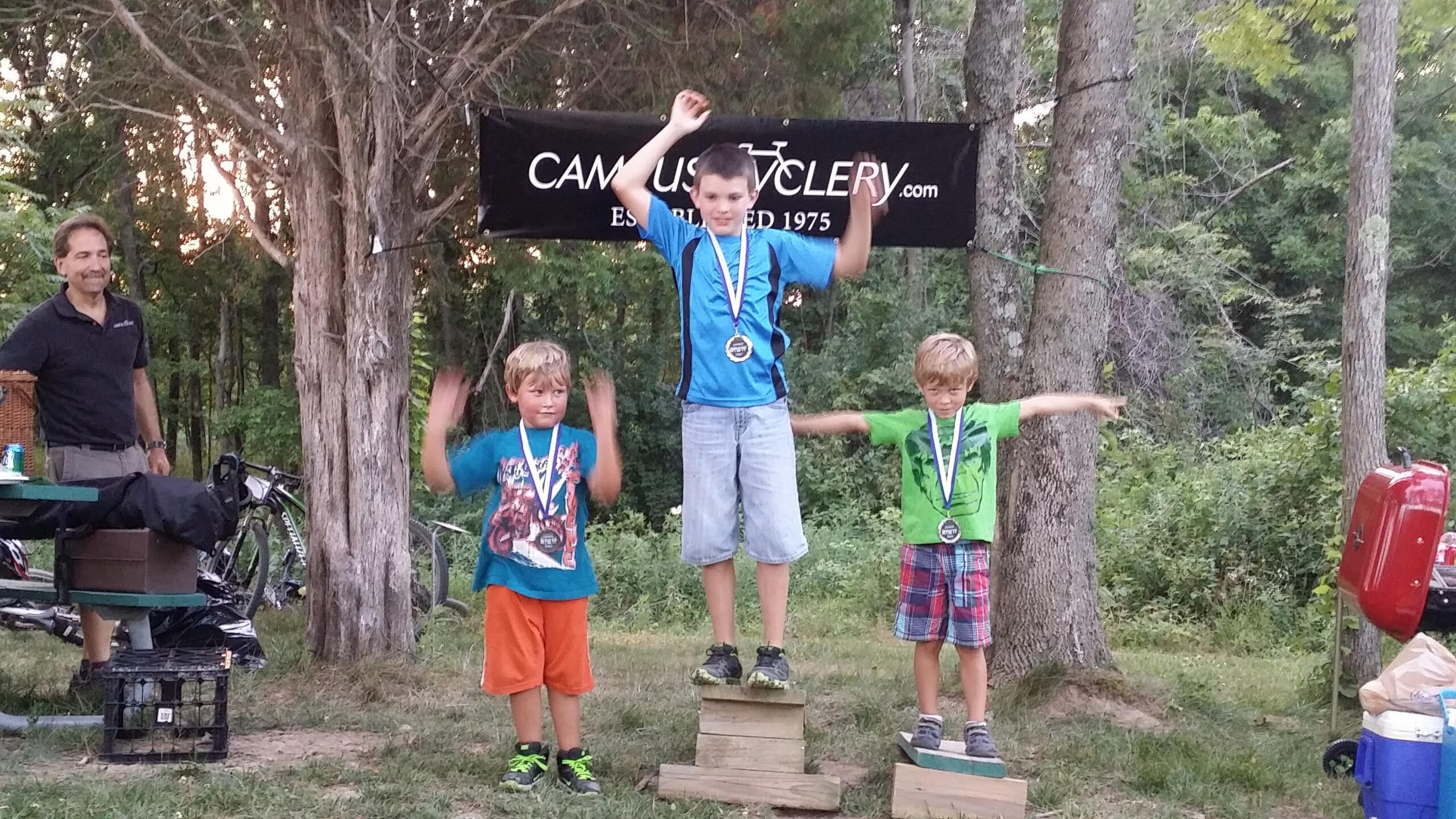 Three young boys stand on wooden podiums, proudly displaying their medals after an award ceremony. The boy in the center, wearing a blue shirt, raises his arms in celebration, while the other two boys, one in a teal shirt and the other in a green shirt with plaid shorts, smile next to him. In the background, a man observes, and several bicycles can be seen nearby amidst a forested setting, with a banner that reads "Campus Cyclery" visible above. England Idlewild Mountain Biking Park mountain bike trail.
