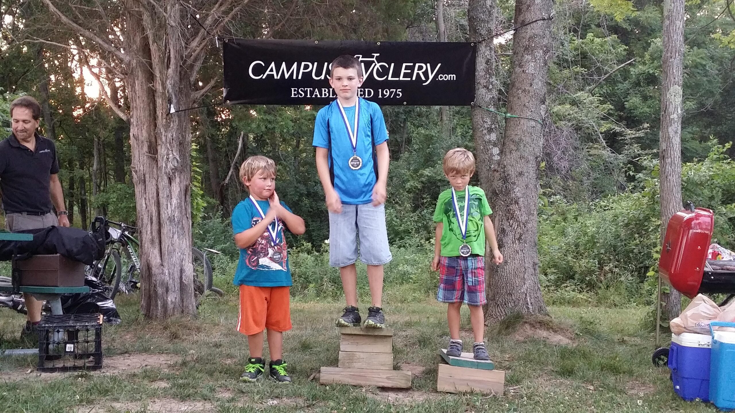 Three young boys stand on podiums, celebrating their achievements at a cycling event. The boy in the middle, wearing a blue shirt and holding a medal, stands confidently on the top tier. The boy to his left, wearing a blue and orange outfit, looks thoughtful, while the boy on the right, dressed in green and plaid, stands calmly. In the background, there is a "Campus Cyclery" banner and trees, with additional equipment visible nearby. England Idlewild Mountain Biking Park mountain bike trail.