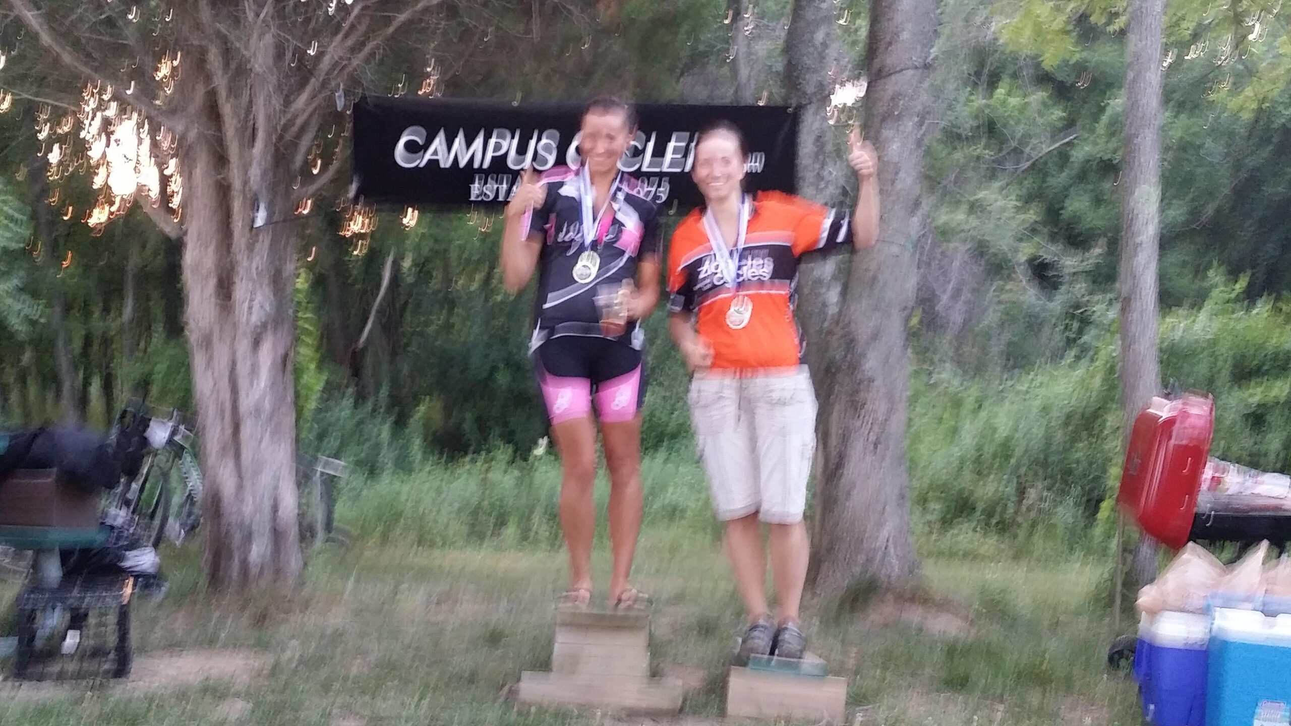 Two participants celebrate on a podium, wearing medals and colorful cycling jerseys. One is in a black jersey with pink accents, and the other is in an orange jersey. They are both smiling and giving thumbs up. The backdrop features a banner that says "CAMPUS CYCLES," and there are trees and a grassy area around them. England Idlewild Mountain Biking Park mountain bike trail.