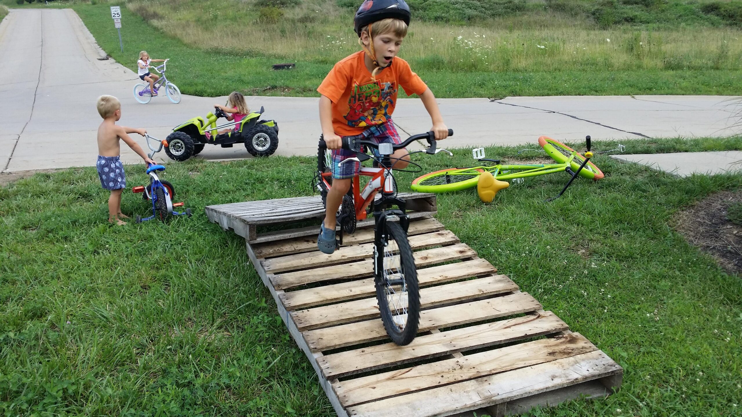A young boy rides a orange bike over a wooden ramp, wearing a helmet and an enthusiastic expression. In the background, another child is riding a small bicycle, while a girl drives a toy vehicle nearby. A second tricycle lies on the grass next to the boy, and other toys are scattered around the scene. The setting appears to be a grassy area next to a road. England Idlewild Mountain Biking Park mountain bike trail.