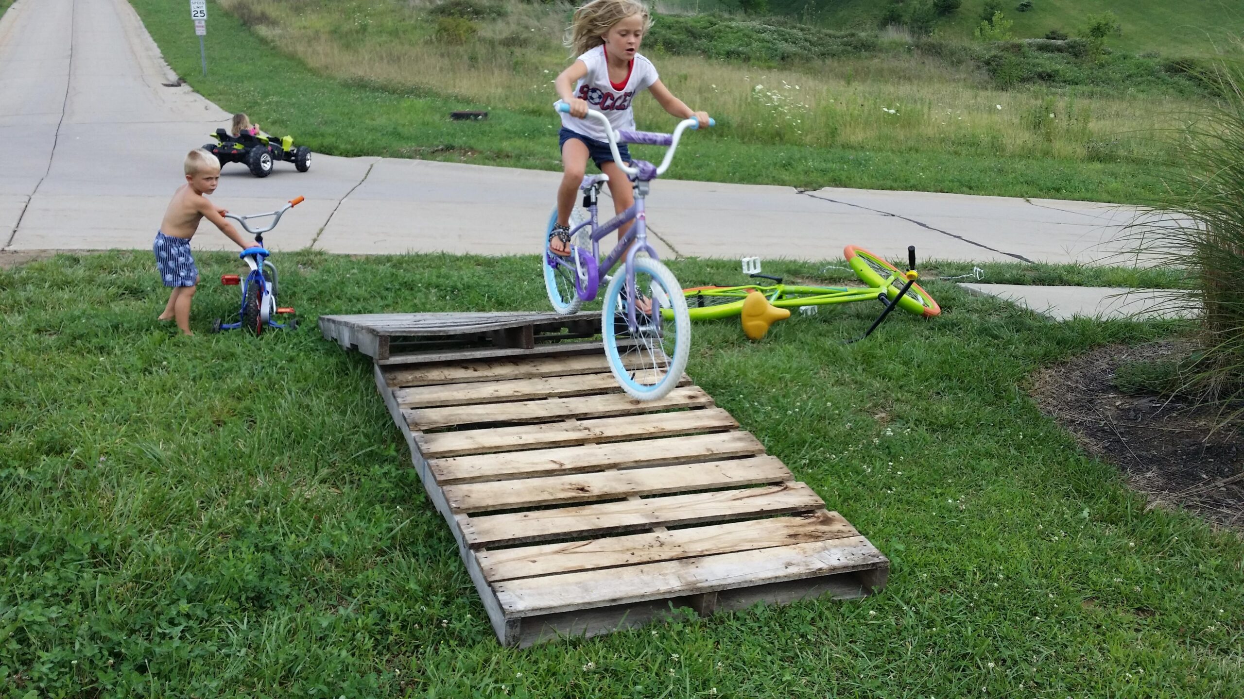 A young girl jumps off a wooden ramp on her bicycle in a grassy area, while a younger boy stands nearby with a blue tricycle. In the background, a child is seen riding a go-kart on the pavement. A colorful bicycle is lying on the grass next to the ramp. England Idlewild Mountain Biking Park mountain bike trail.