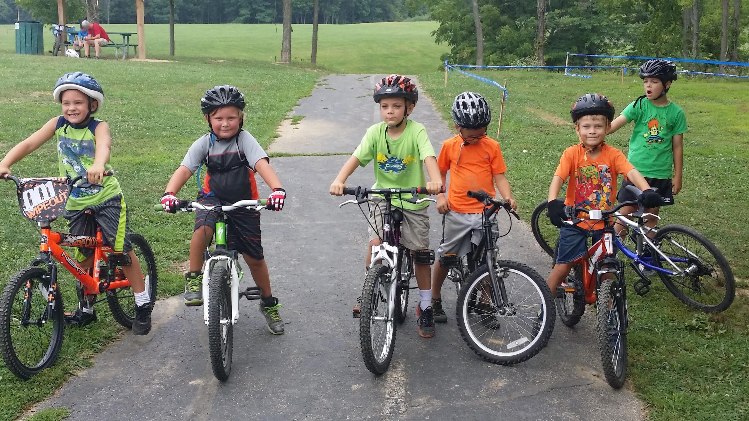 A group of five young boys wearing helmets and riding bicycles on a paved path in a park. They are smiling and posing for the camera, with a grassy area and trees in the background. The boys are dressed in colorful t-shirts and shorts, and various types of bikes can be seen. England Idlewild Mountain Biking Park mountain bike trail.