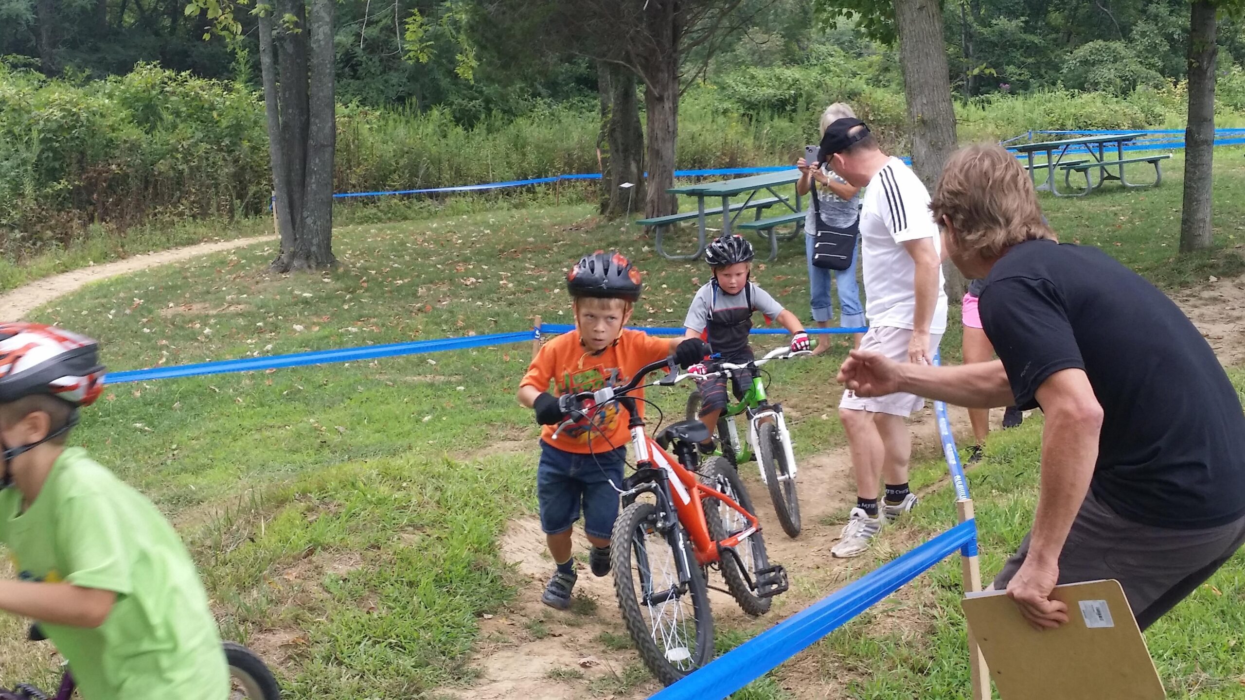 A group of young children participate in a bike race, with two boys in the foreground pushing their bikes along a grassy path marked with blue tape. One boy wears an orange shirt and a helmet, while the other is in a gray shirt. Nearby, adults are cheering and monitoring the event. Trees and picnic tables can be seen in the background, creating a natural setting for the race. England Idlewild Mountain Biking Park mountain bike trail.