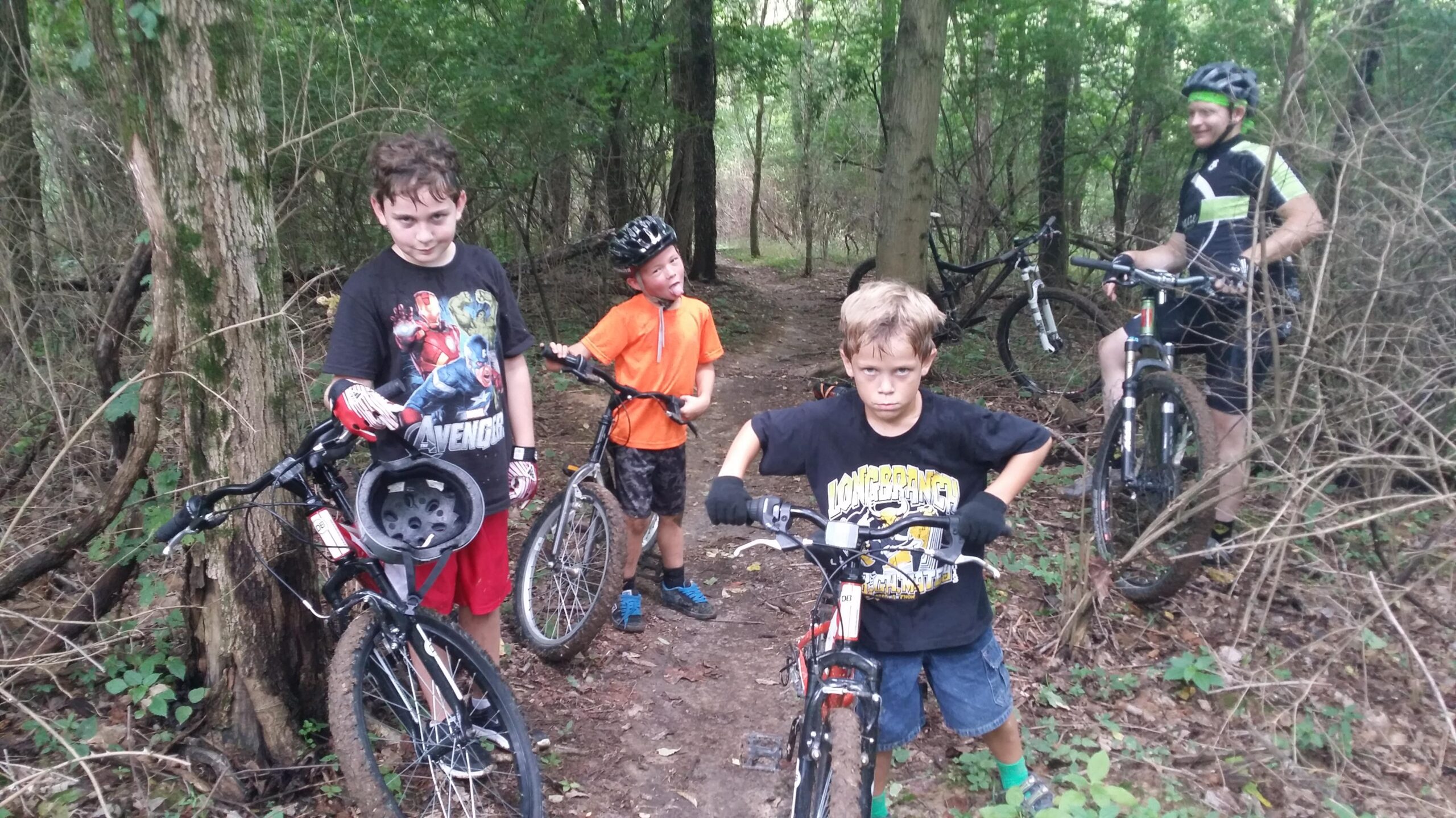 A group of three boys and one adult man are standing in a wooded area, surrounded by trees and vegetation. The boys are positioned near their bicycles; one boy is wearing a black Avengers shirt and red shorts, another is in an orange shirt and black shorts, and the third boy is in a black shirt with yellow graphics. The adult man, dressed in cycling gear, is positioned further back with his bike. The atmosphere is casual, and the boys appear playful, with one sticking out his tongue. England Idlewild Mountain Biking Park mountain bike trail.