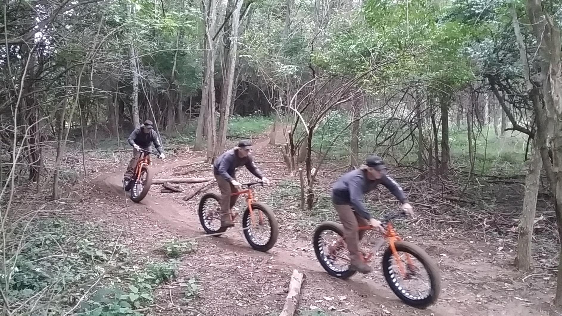 Three mountain bikers riding on a dirt trail through a wooded area, surrounded by trees and greenery. Each biker is on a fat bike, navigating a slight incline. One rider is slightly ahead while the others are in motion behind them. England Idlewild Mountain Biking Park mountain bike trail.