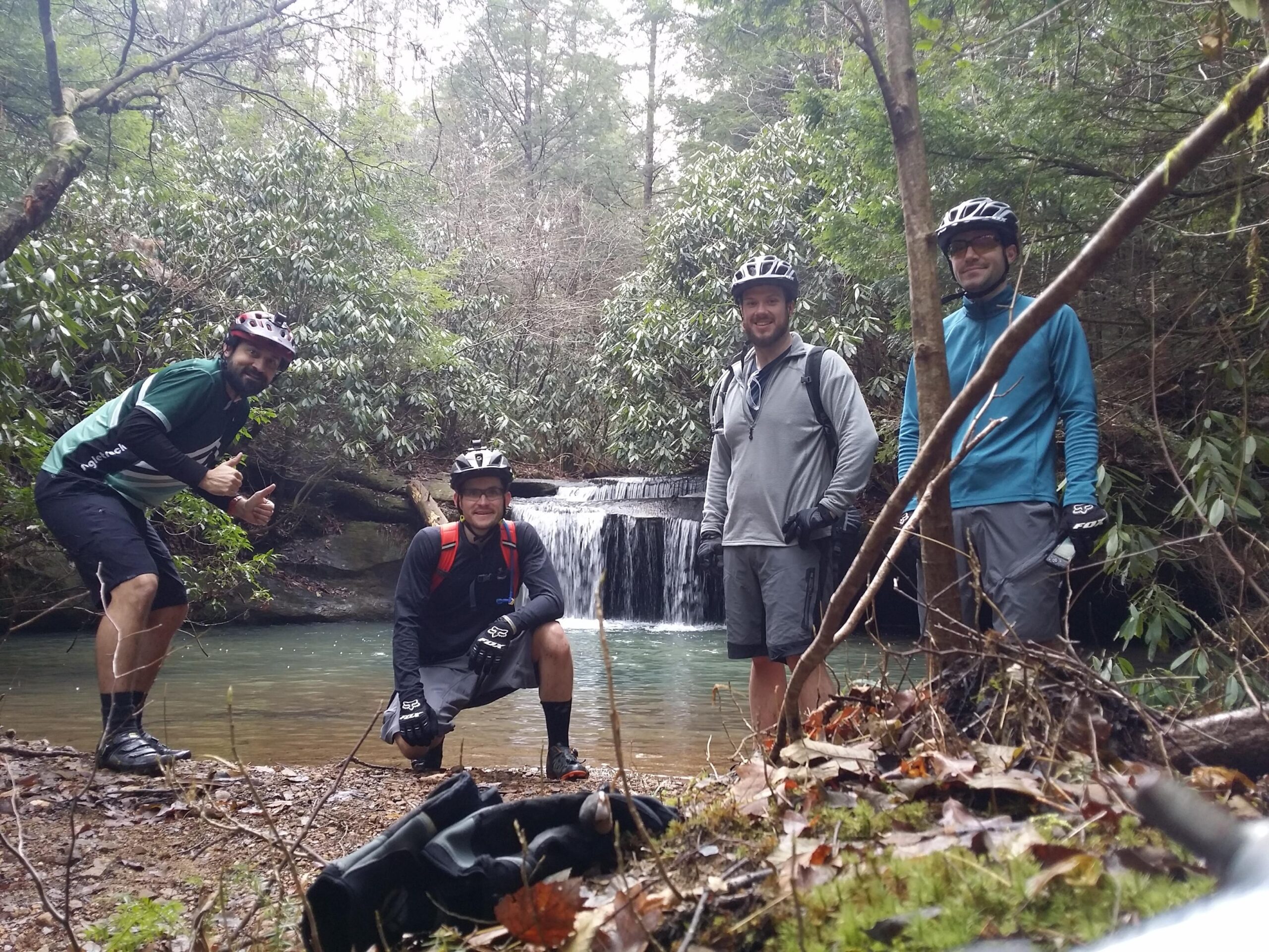 Four mountain bikers posing near a small waterfall in a forested area, surrounded by greenery and fallen leaves. Two of the bikers are giving thumbs up, while others smile, all wearing helmets and biking gear. Cane Creek (sheltowee Trace Trail) mountain bike trail.