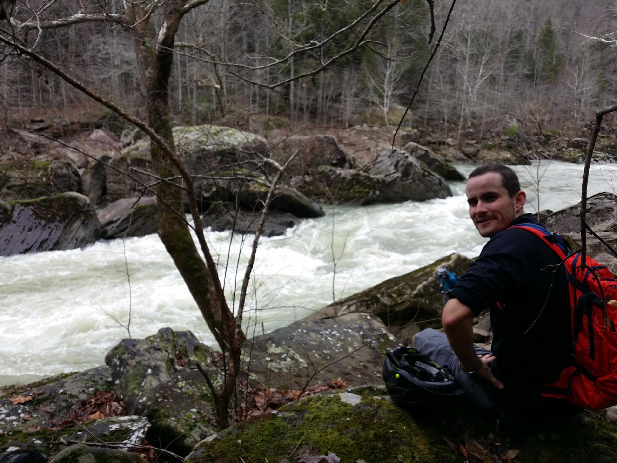 A person sitting on a rocky riverbank, smiling while looking over their shoulder towards the camera. The background features a rushing river surrounded by trees and boulders, indicating a natural outdoor setting. The individual is wearing a black shirt and has a red backpack beside them. Cane Creek (sheltowee Trace Trail) mountain bike trail.