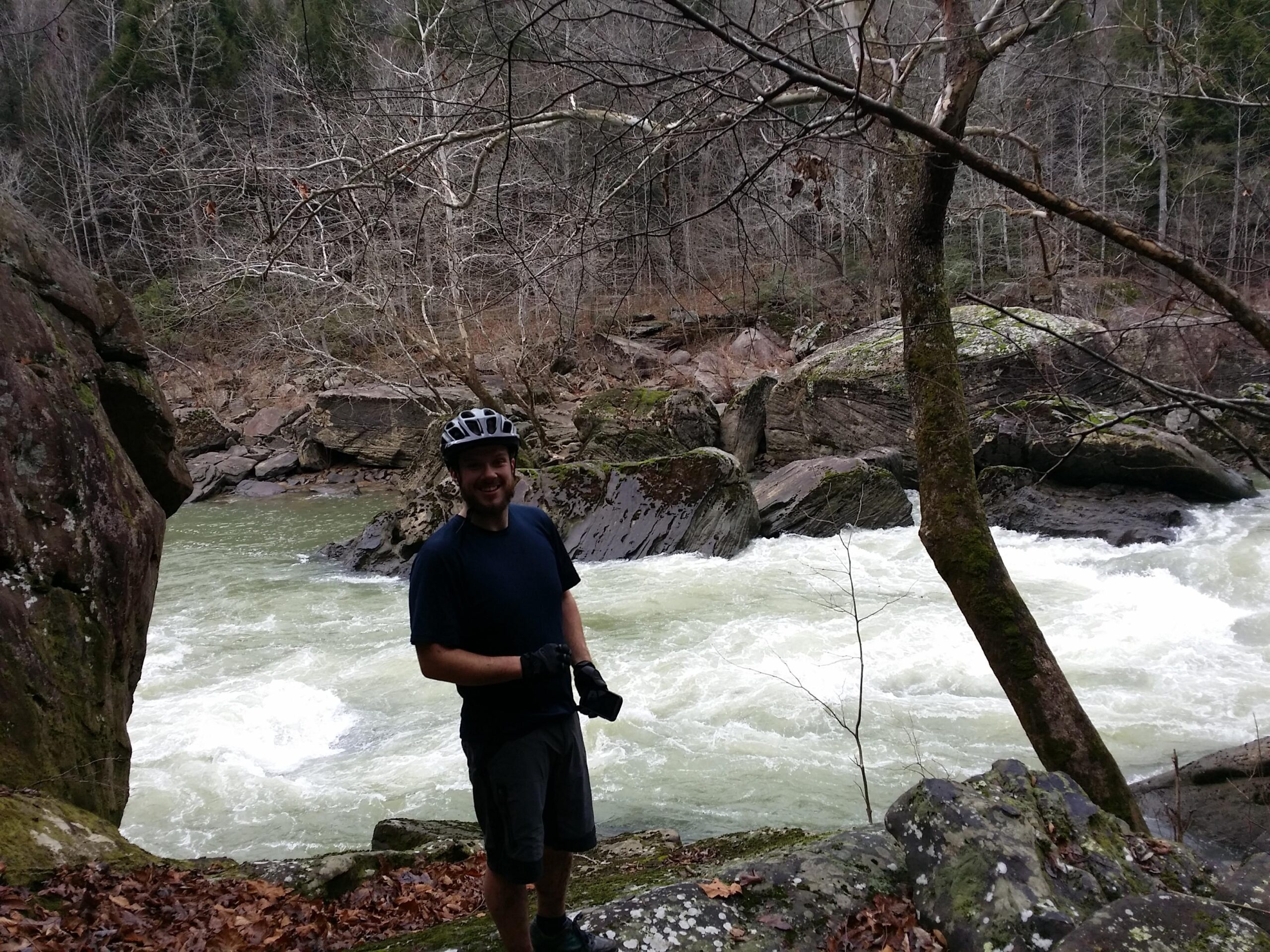 A man wearing a helmet and gloves stands on the rocky bank of a river, smiling at the camera. The river is flowing rapidly, surrounded by moss-covered rocks and bare trees in a natural landscape. The scene reflects a rugged outdoor environment, suggesting a location for activities such as mountain biking or hiking. Cane Creek (sheltowee Trace Trail) mountain bike trail.
