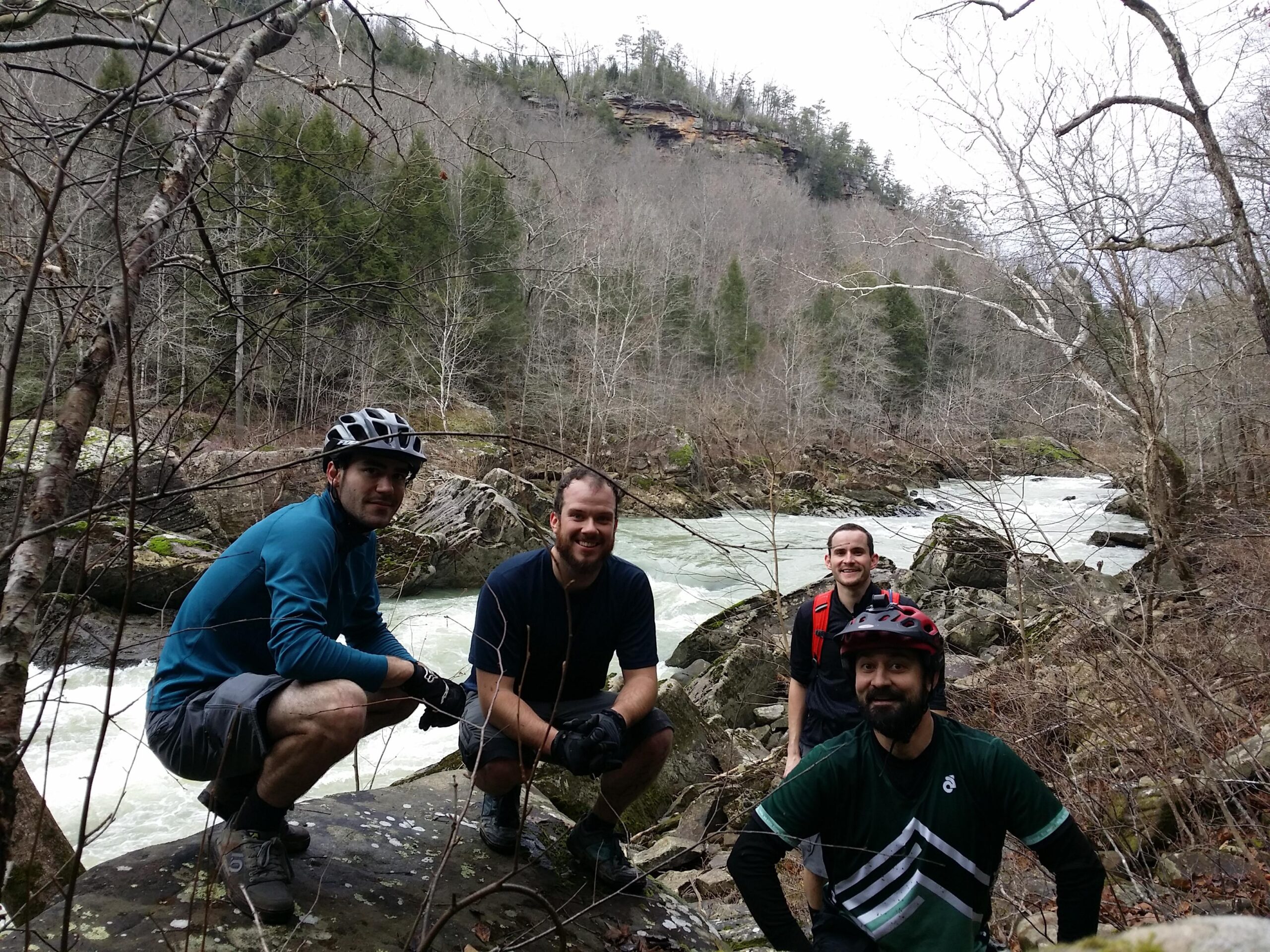 Four men in cycling gear pose for a photo near a riverbank, surrounded by a forest. The background features trees with sparse leaves and rocky terrain, while the river flows swiftly beside them. The atmosphere suggests an outdoor adventure, likely after biking or hiking. Cane Creek (sheltowee Trace Trail) mountain bike trail.