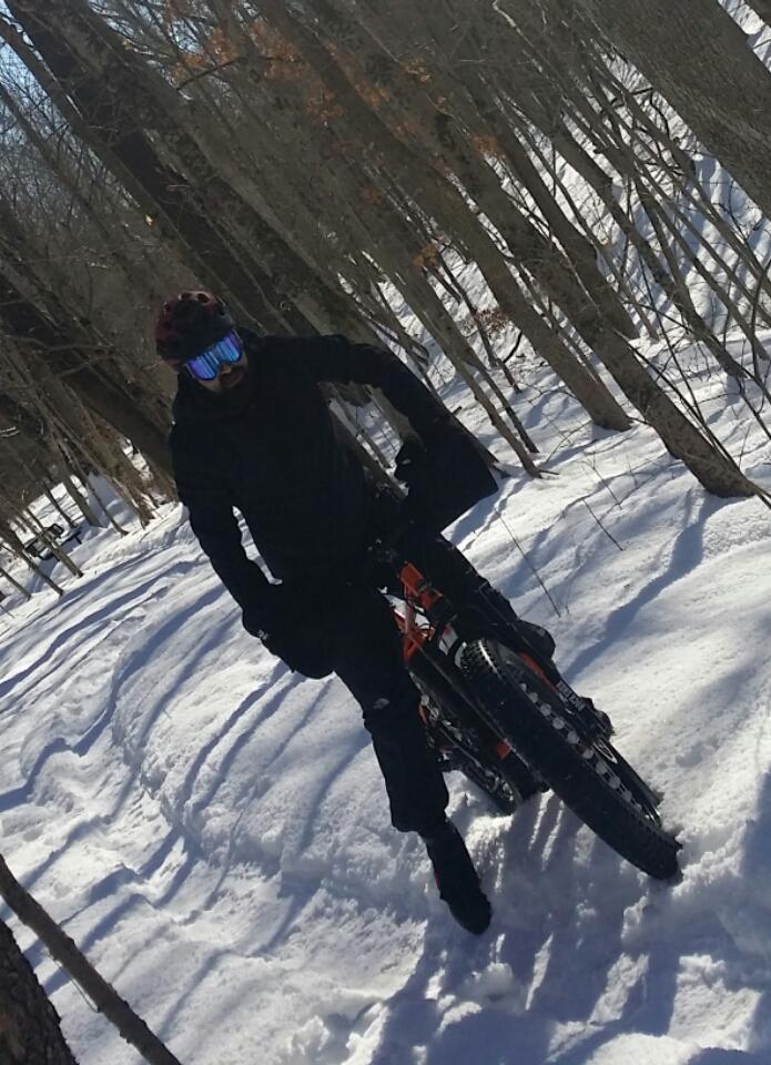 A person in black outdoor gear and a helmet is riding a mountain bike through a snowy forest trail. The bike features wide tires designed for snow, and the surrounding trees are bare, indicating a winter setting. The individual is leaning slightly forward, navigating the snowy terrain. England Idlewild Mountain Biking Park mountain bike trail.