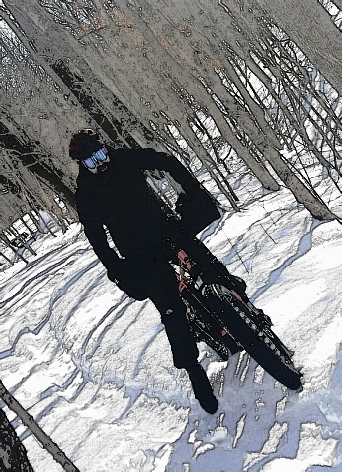 A person dressed in black winter clothing and goggles is riding a bicycle through a snowy forest, surrounded by bare trees. The scene captures the winter landscape with visible tire tracks in the snow. England Idlewild Mountain Biking Park mountain bike trail.