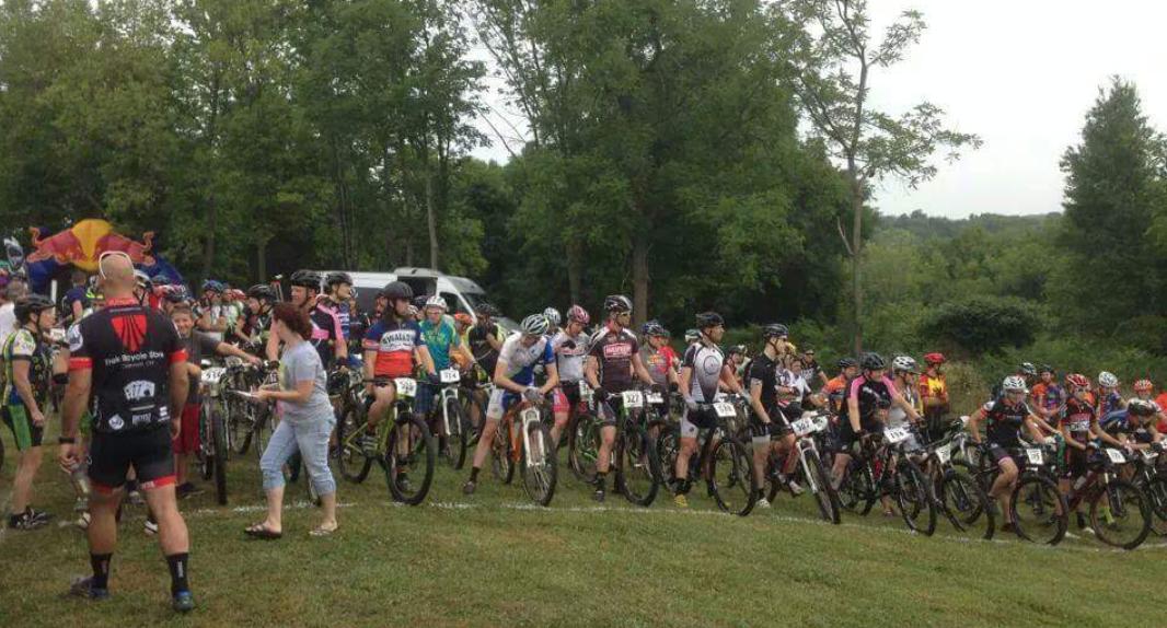 A large group of cyclists, wearing various colorful jerseys and helmets, gather on a grassy area before an event. Some riders are holding their mountain bikes while others chat. The background features trees and a few vehicles, creating a lively atmosphere for the cycling event. England Idlewild Mountain Biking Park mountain bike trail.