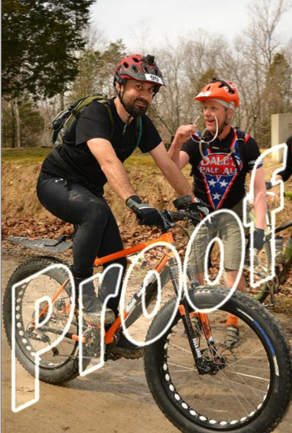 Two mountain bikers riding on a dirt trail. One rider is laughing and gesturing while wearing an orange helmet and a colorful cycling shirt. The other rider has a black helmet and is wearing a black shirt and shorts, focused on the trail ahead. The image has the word "Proof" overlayed in large, translucent letters. In the background, trees and a blurred path can be seen. Hickory Ridge mountain bike trail.