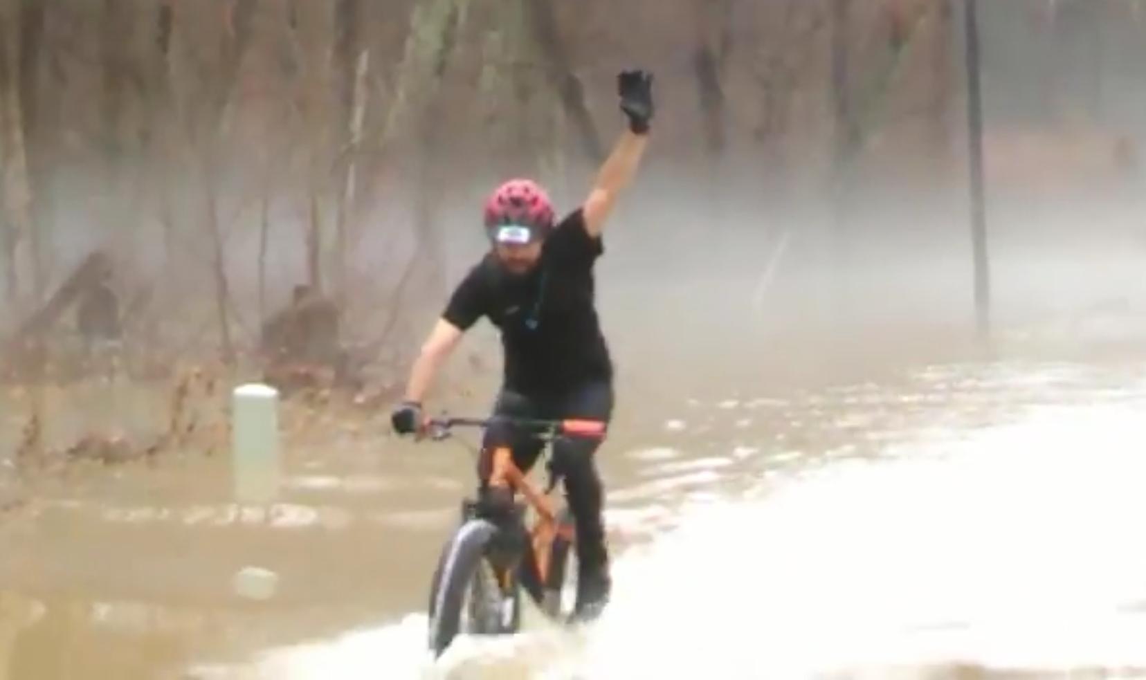A person riding a mountain bike through flooded waters, raising one hand in the air. The background features trees and mist, indicating a foggy atmosphere. The cyclist is wearing a helmet, gloves, and casual athletic clothing. Hickory Ridge mountain bike trail.