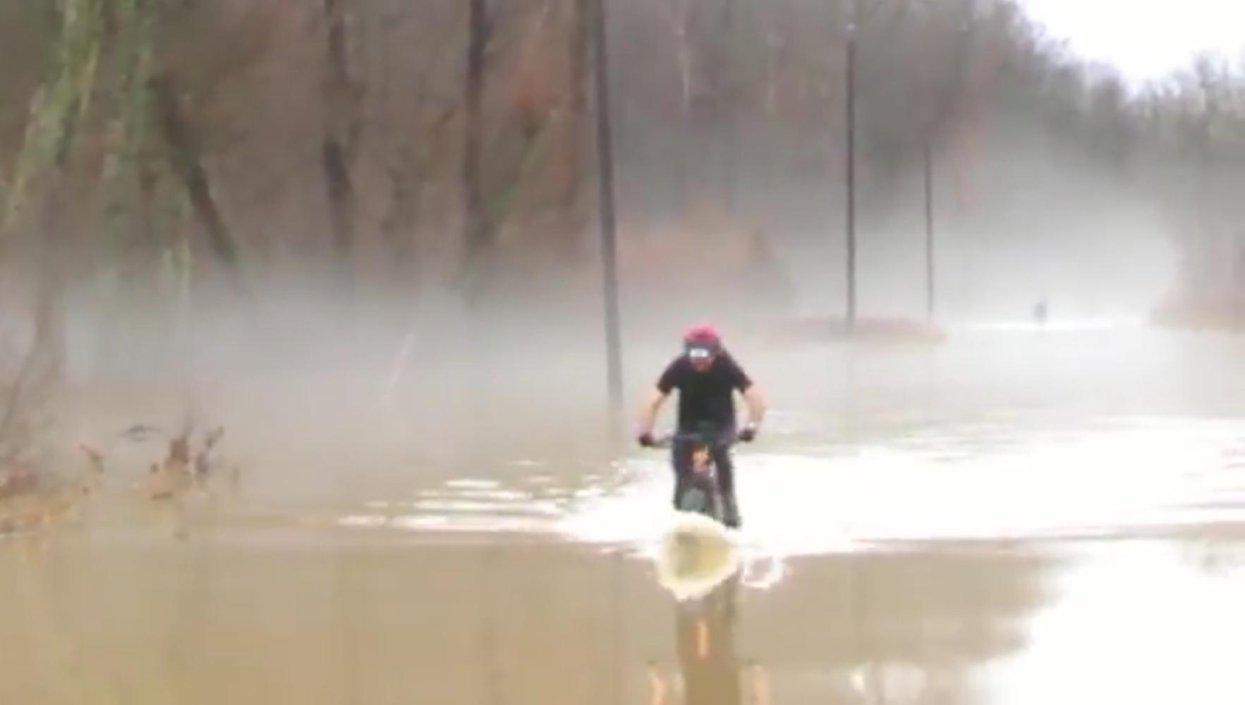 A person riding a bicycle through a flooded area, with water covering the ground and mist rising from the surface. Trees and power poles are visible in the background, indicating a natural setting affected by flooding. Hickory Ridge mountain bike trail.