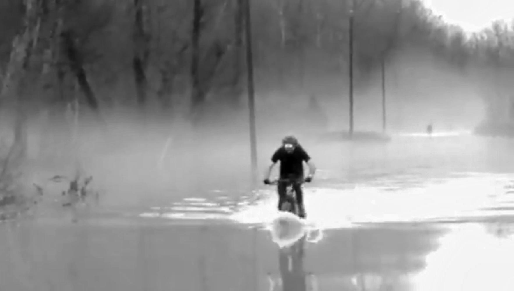 A person riding a bicycle through a flooded area, surrounded by fog and bare trees. The scene is monochrome, emphasizing the misty atmosphere and waterlogged landscape. Hickory Ridge mountain bike trail.