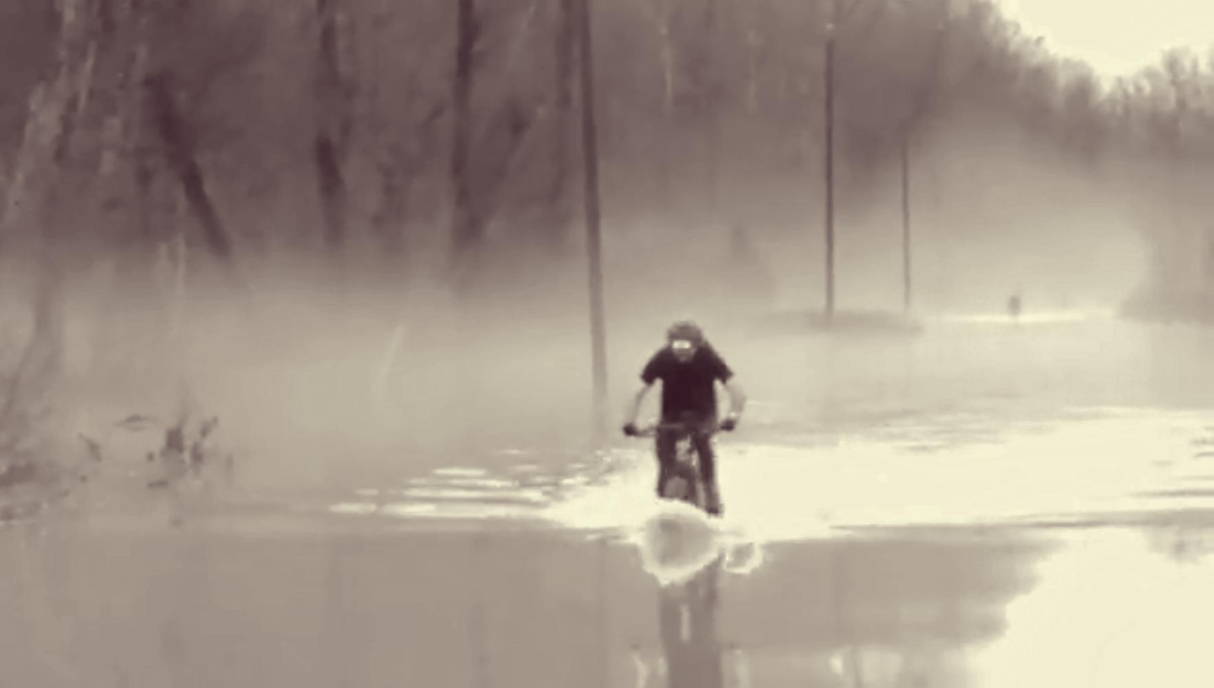 A person riding a mountain bike through flooded water, creating splashes, with trees and poles visible in a misty background. The image is in black and white, giving it a dramatic and atmospheric feel. Hickory Ridge mountain bike trail.