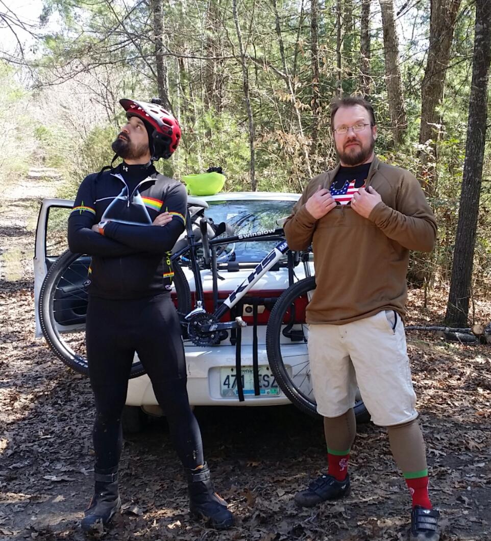 Two men stand confidently in front of a parked vehicle on a wooded trail. One man, wearing a black cycling outfit and a red helmet, looks upwards with his arms crossed. The other man, dressed in a brown long-sleeve shirt and beige shorts, playfully lifts his shirt to reveal an American flag design underneath. A bicycle is secured on the back of the vehicle, and the scene takes place in a natural, leafy environment. Big South Fork mountain bike trail.