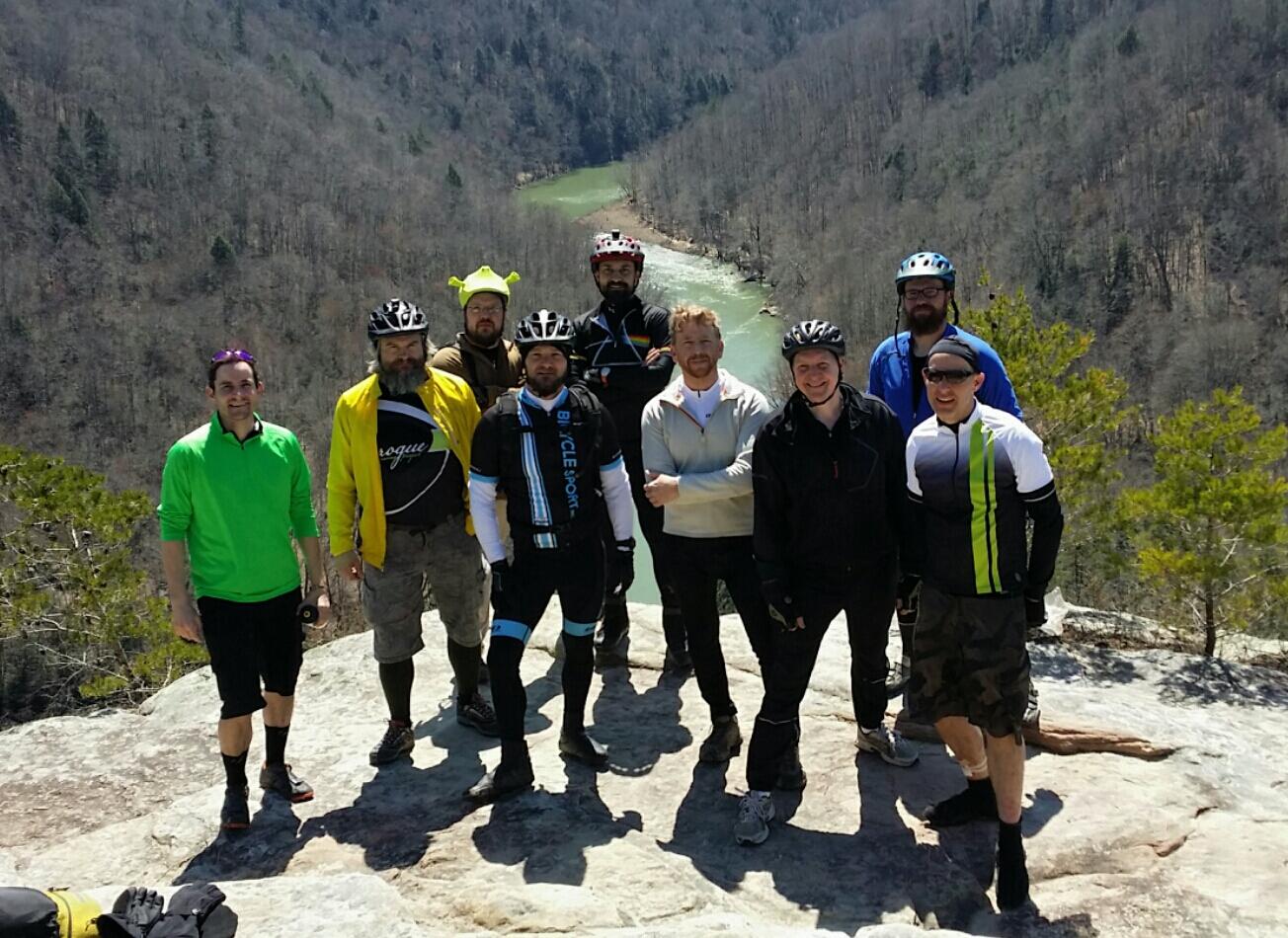 A group of nine men, dressed in cycling gear and helmets, stand on a rocky outcrop with a scenic view of a river and wooded landscape in the background. The men display a variety of expressions and poses, some with smiles, while the sun shines down on the outdoor setting. Big South Fork mountain bike trail.