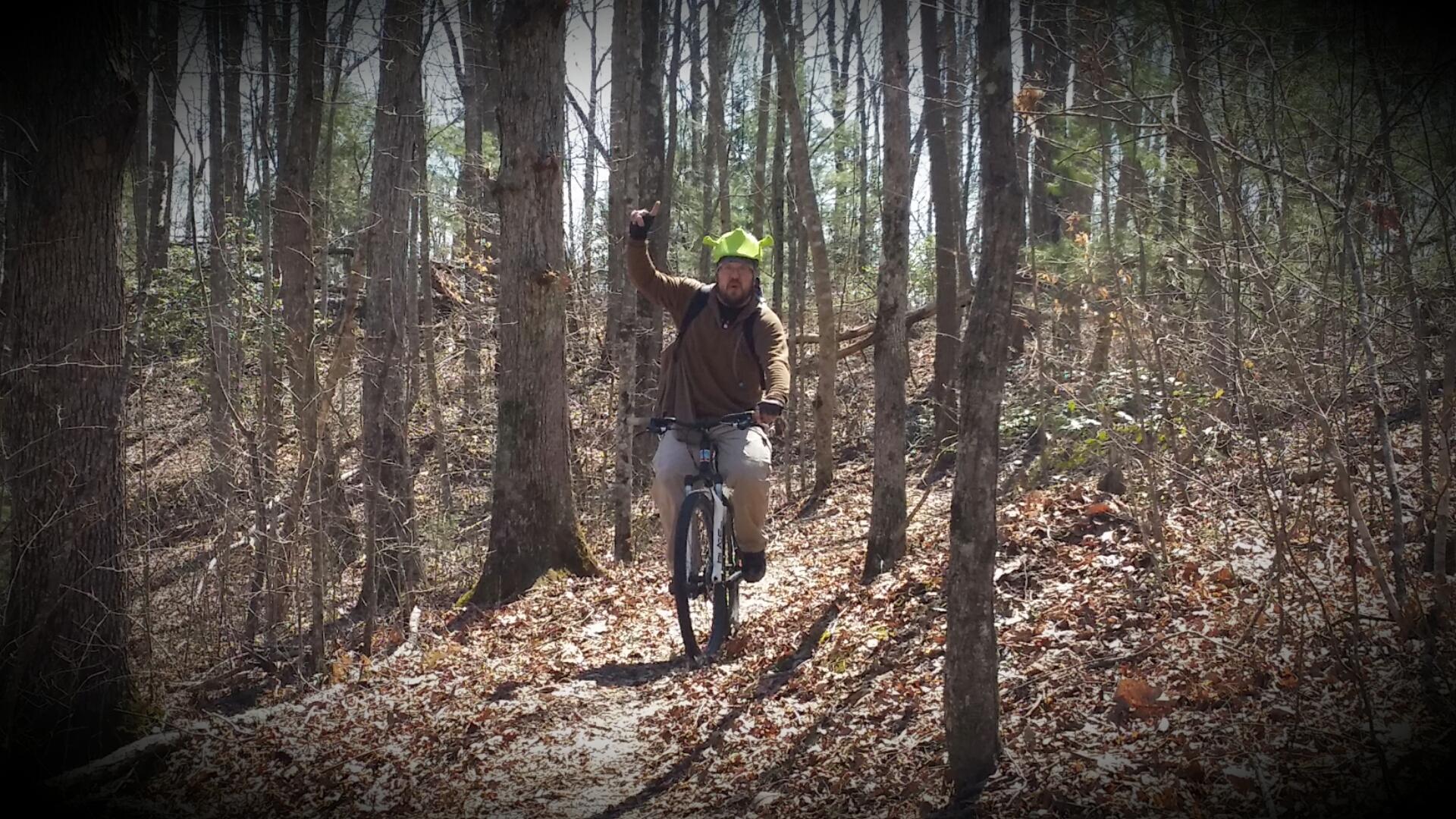 A person riding a mountain bike on a wooded trail, raising one arm in a playful gesture. The rider is wearing a green, novelty hat and dressed in casual outdoor clothing. Surrounding trees have sparse leaves, indicating an early spring or late winter setting. The ground is covered with fallen leaves. Big South Fork mountain bike trail.