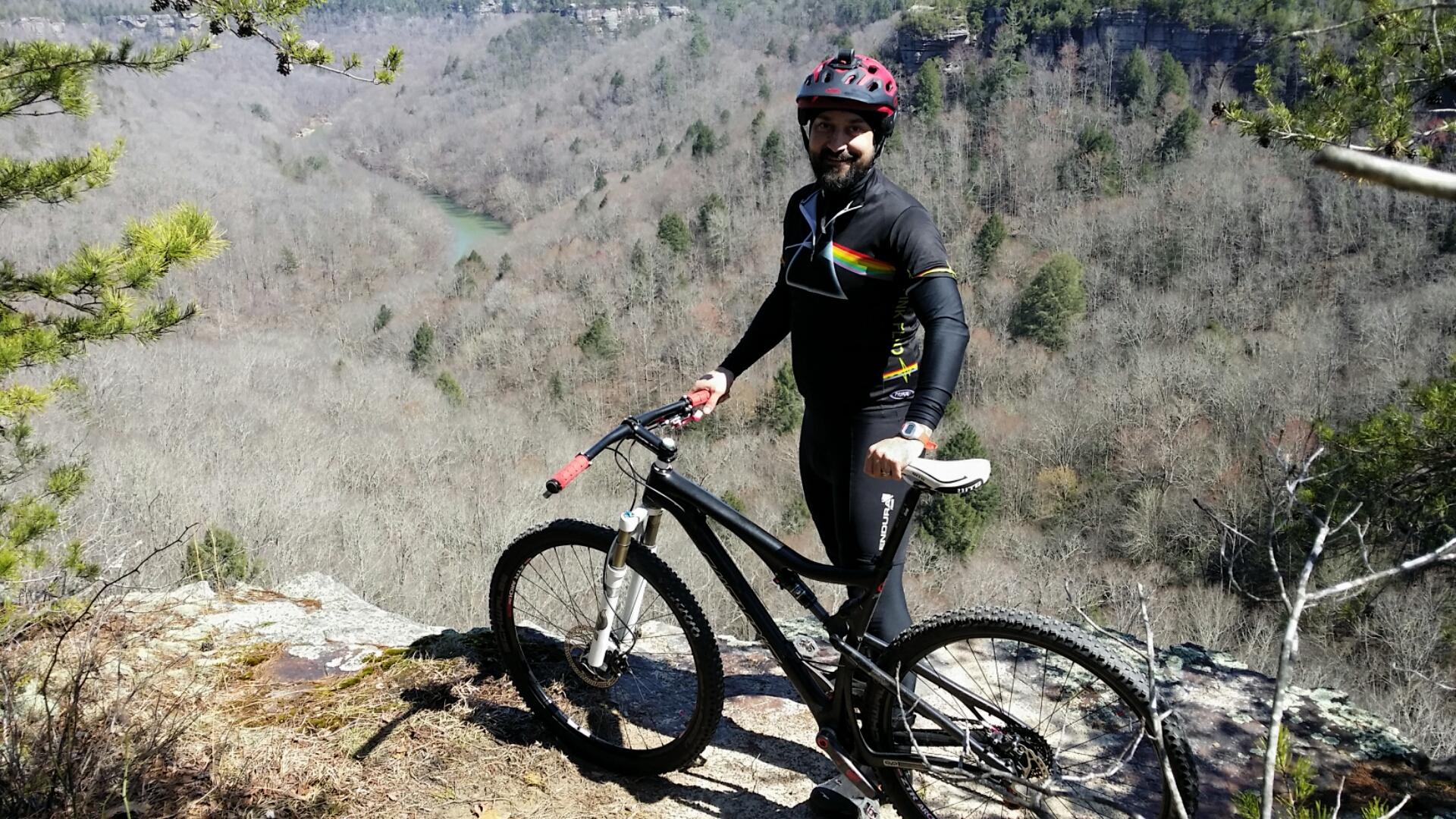 A person wearing a helmet and cycling gear stands next to a mountain bike on a rocky overlook, surrounded by a forest of bare trees and a winding river in the background. The scene is bright and sunny, showcasing a beautiful outdoor landscape. Big South Fork mountain bike trail.