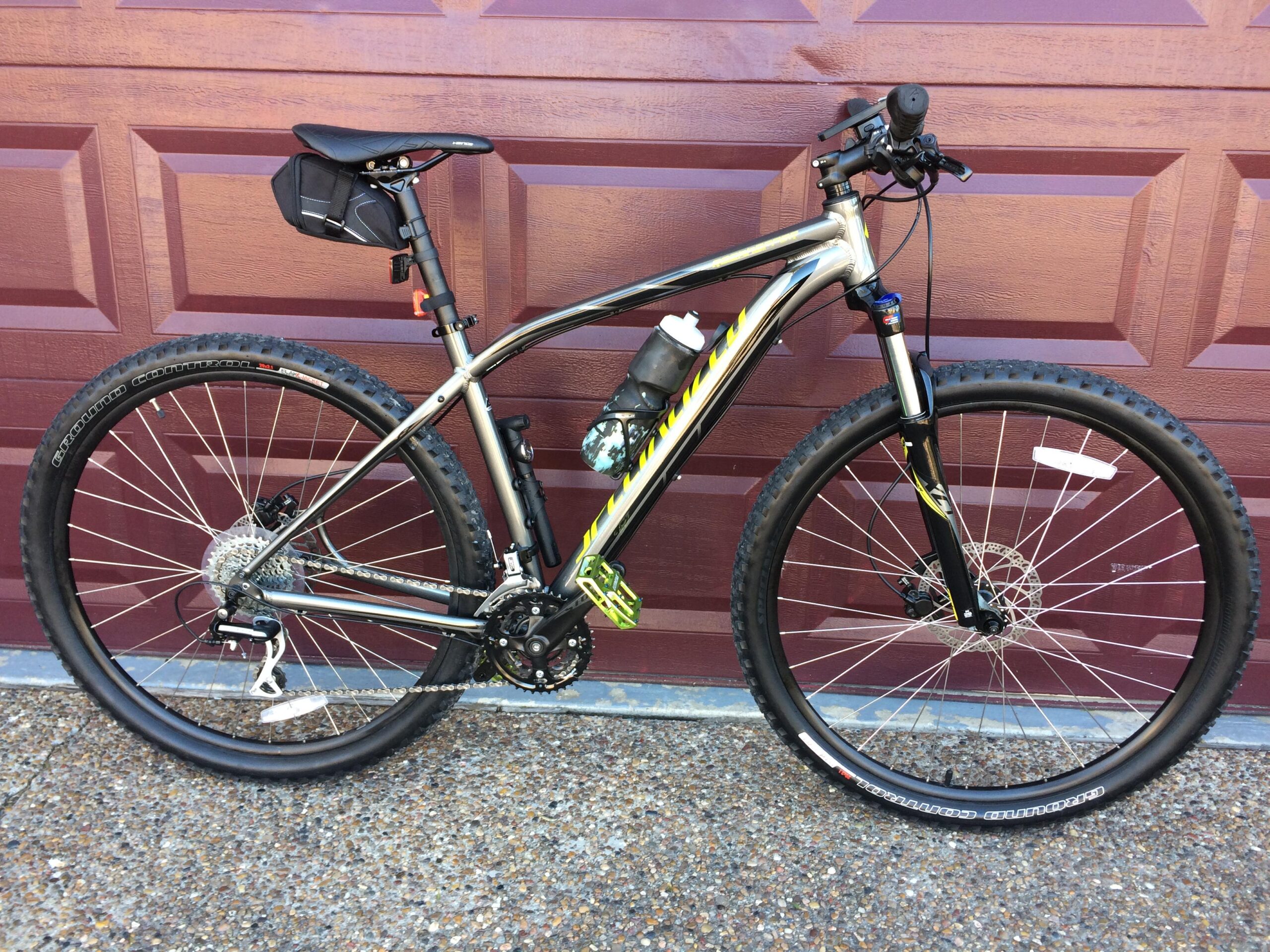 Specialized Rockhopper 29: A mountain bike with a silver and black frame is leaning against a maroon garage door. The bike features thick tires, a water bottle mounted on the frame, and a small black saddle bag attached to the seat. The bike has a modern design with visible gears and disc brakes.
