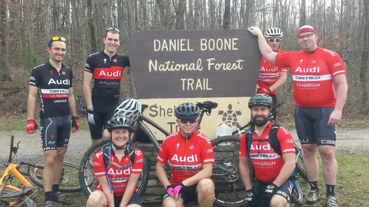 A group of eight cyclists in athletic apparel, posing in front of a sign that reads "Daniel Boone National Forest Trail." The scene is set in a wooded area, with trees in the background, and several bicycles are leaning against the sign. The cyclists are smiling and appear to be enjoying their outdoor adventure.