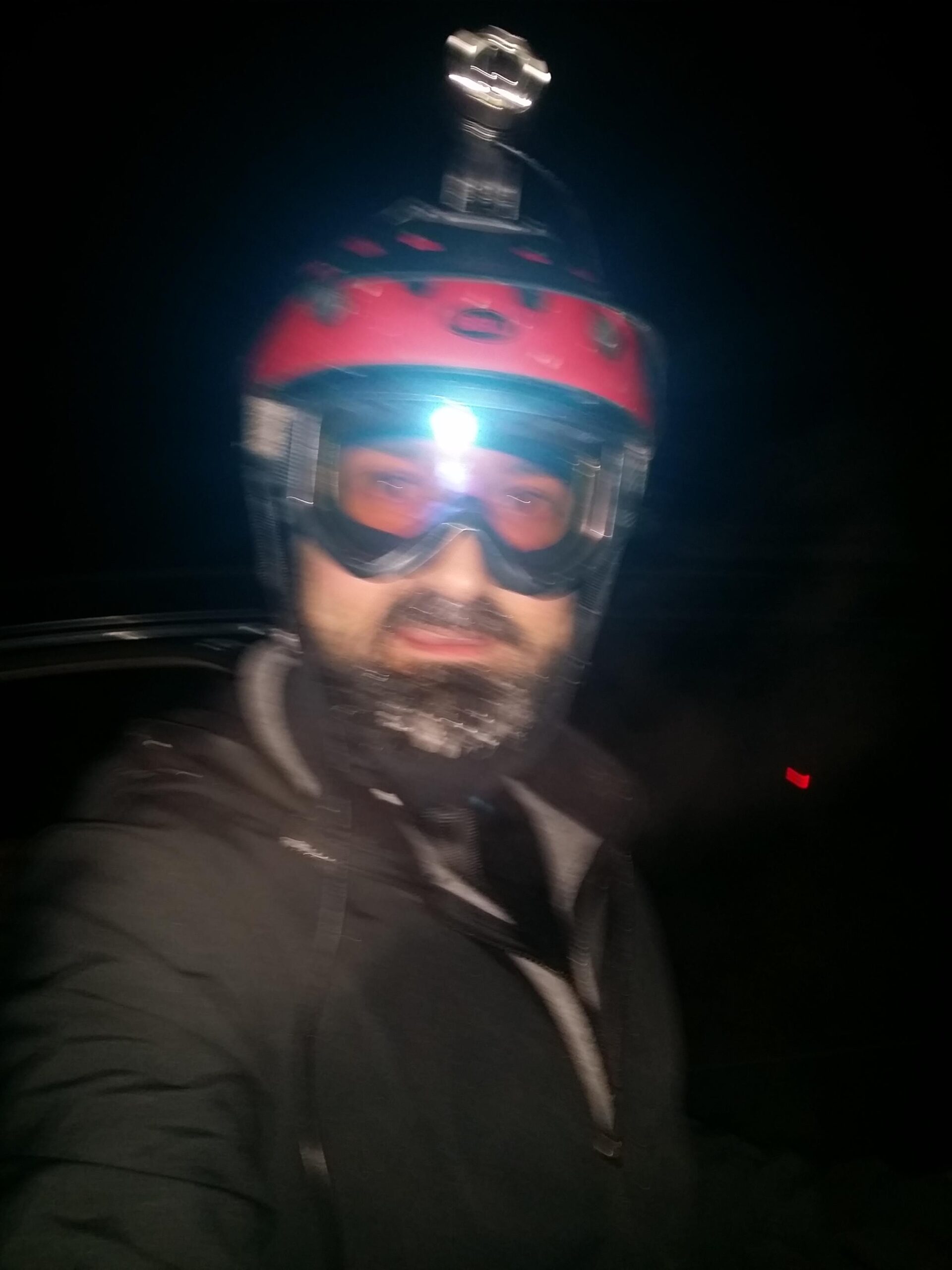 A person wearing a red helmet with a headlamp and goggles, dressed in a black jacket, standing outdoors at night. Snow or frost is visible on their beard, indicating cold weather conditions. The background is dark, suggesting a nighttime environment. England Idlewild Mountain Biking Park mountain bike trail.