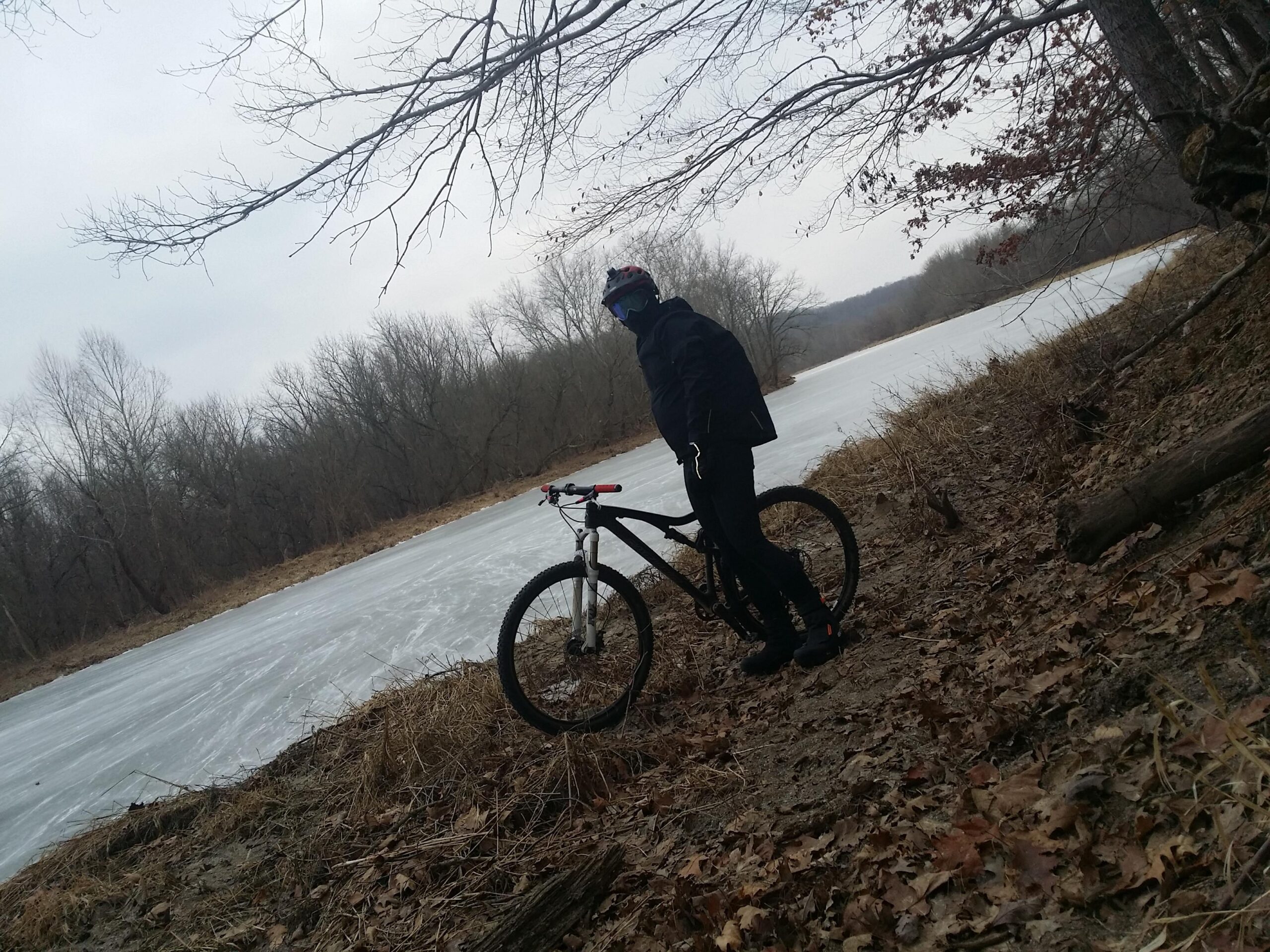 A person in winter attire stands next to a mountain bike on the edge of a frozen river. The background features bare trees and a cloudy sky, with brown leaves scattered on the ground. Versailles State Park mountain bike trail.