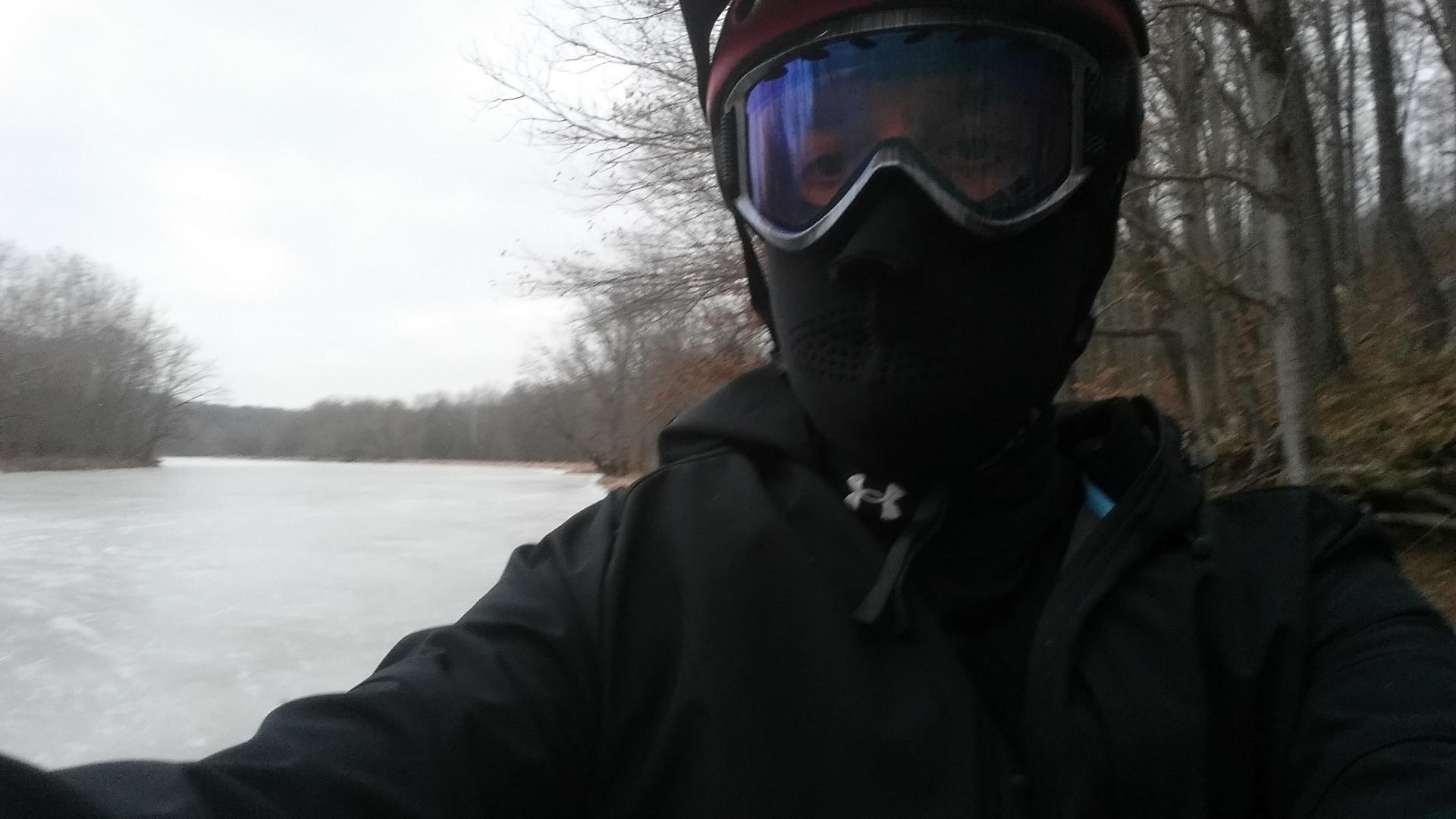 A person dressed warmly in a black mask and goggles takes a selfie by a frozen river. The landscape includes bare trees and a cloudy sky, reflecting a wintry atmosphere. Versailles State Park mountain bike trail.