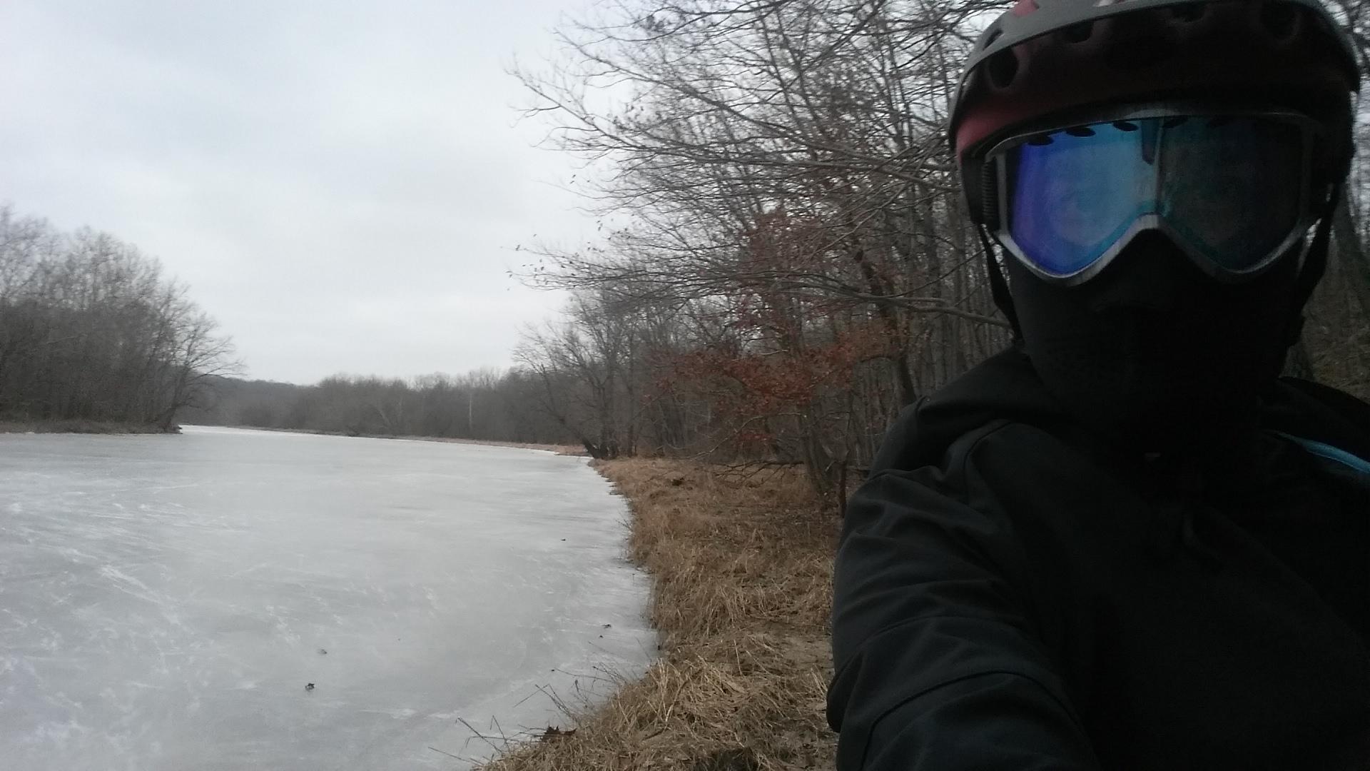 A person wearing a helmet and goggles stands beside a frozen lake, surrounded by bare trees and a grassy shoreline. The sky is overcast, suggesting a cold winter day. Versailles State Park mountain bike trail.