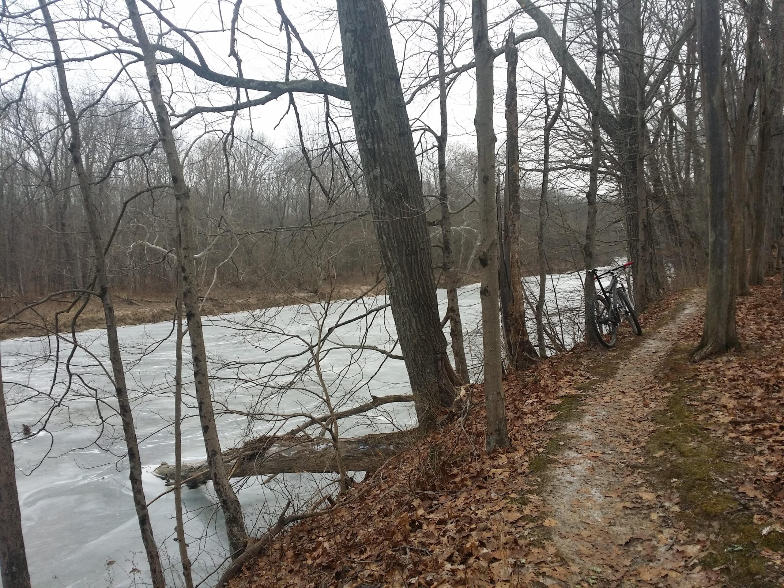 A winter scene along a riverbank featuring a partially frozen river. A winding dirt path runs beside the river, lined with bare trees. A mountain bike leans against one of the trees, and fallen leaves are scattered along the ground. The atmosphere is gray and chilly, suggesting a cold day. Versailles State Park mountain bike trail.