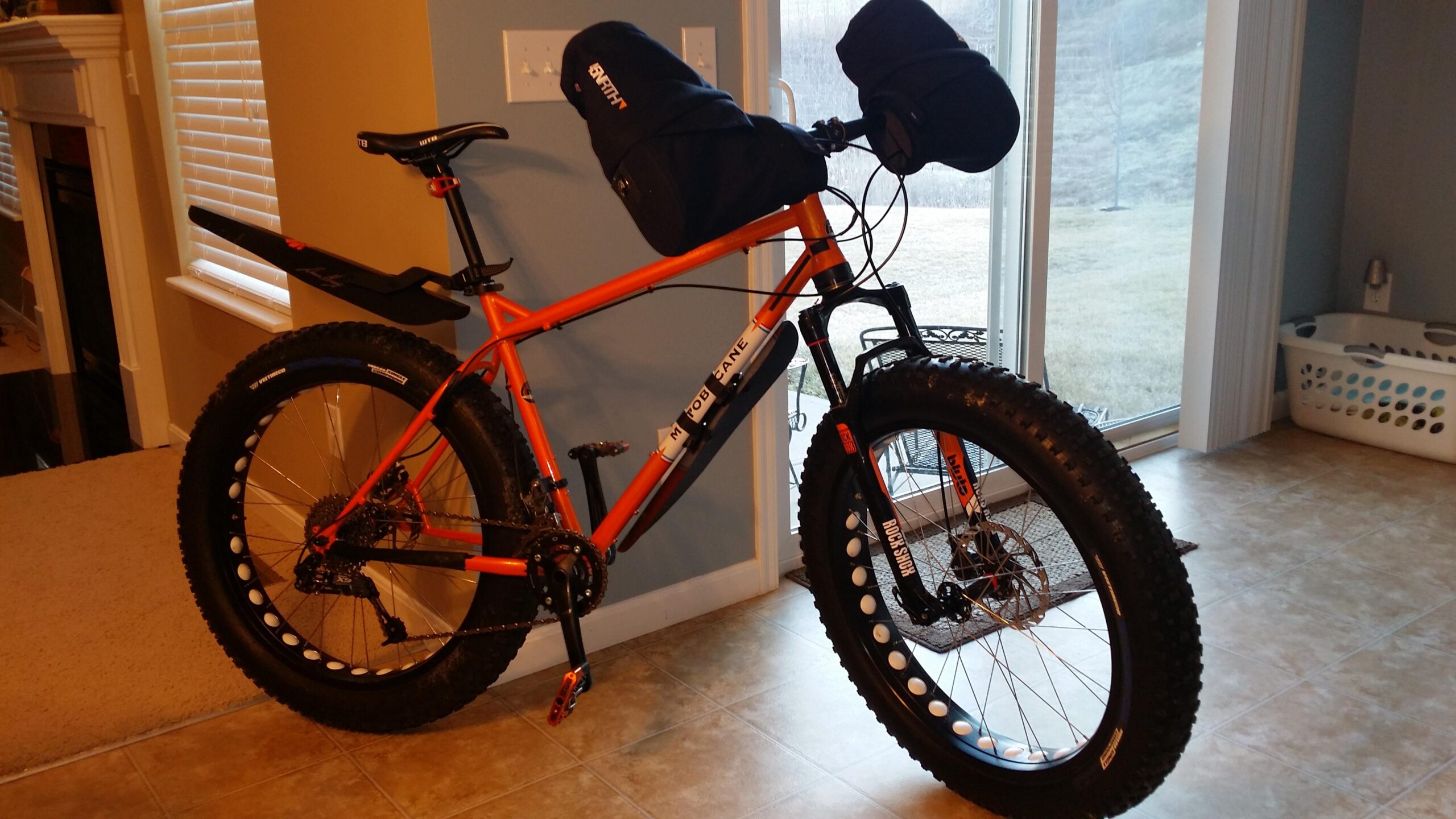 Motobecane Lurch: Alt text: An orange fat tire bike is positioned indoors next to a window, showcasing large, wide tires and a sleek design. The bike features a black saddle bag on the handlebars and a rear rack with a tail light. Sunlight streams in through the window, illuminating the tiled floor and nearby furnishings.