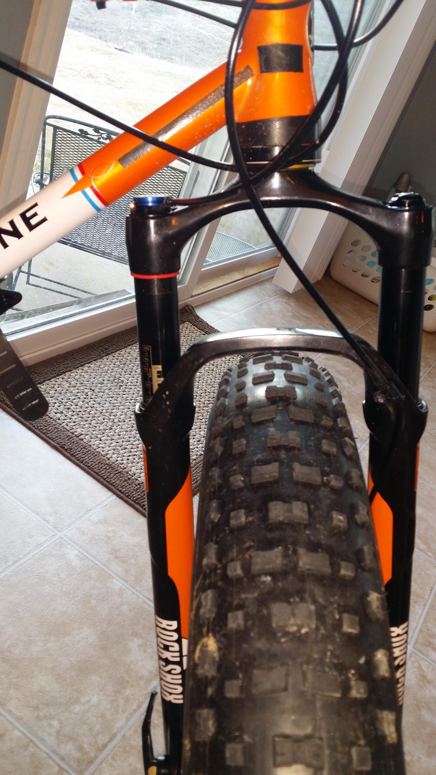 Motobecane Lurch: A close-up view of a mountain bike's front fork and tire, showcasing an orange and black design. The image highlights the fork's suspension components, the tread of the tire, and part of the bike frame, with a blurred background of a patio area.
