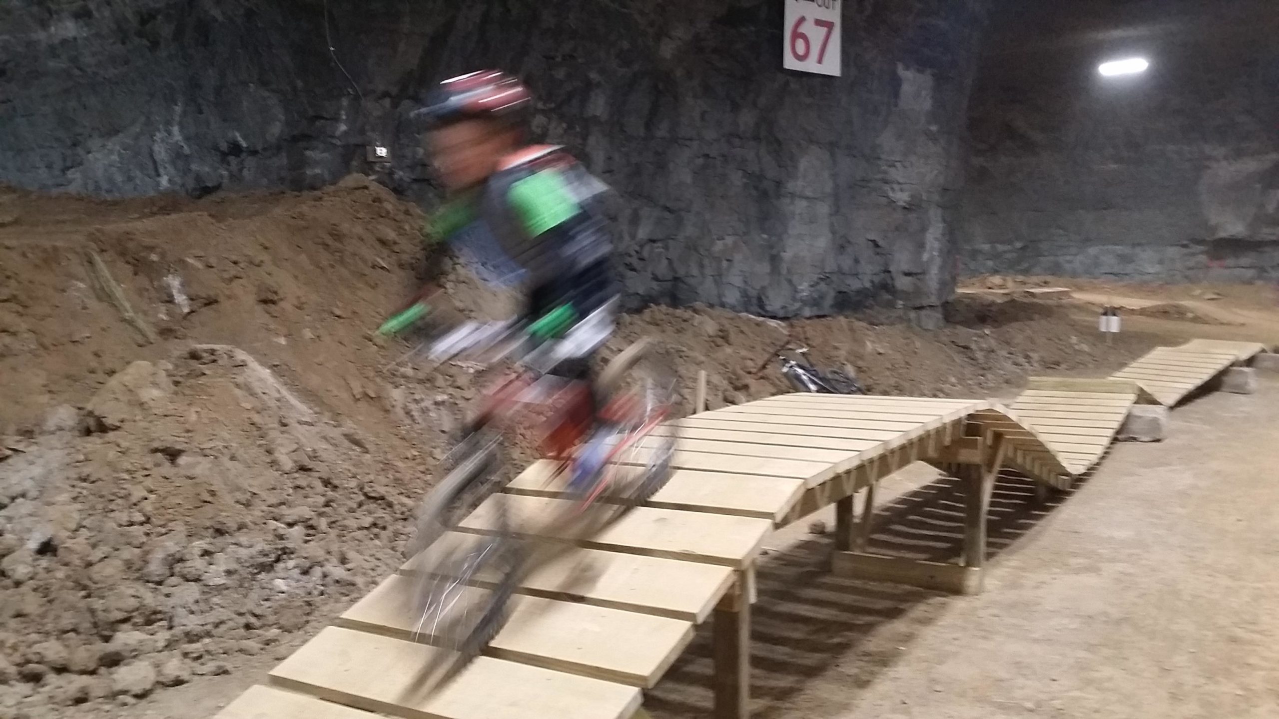A blurred image of a cyclist riding a bike on a wooden ramp in an underground biking facility, with rough terrain and rock walls visible in the background. A sign with the number 67 is attached to the wall above. Louisville Mega Cavern Bike Park mountain bike trail.
