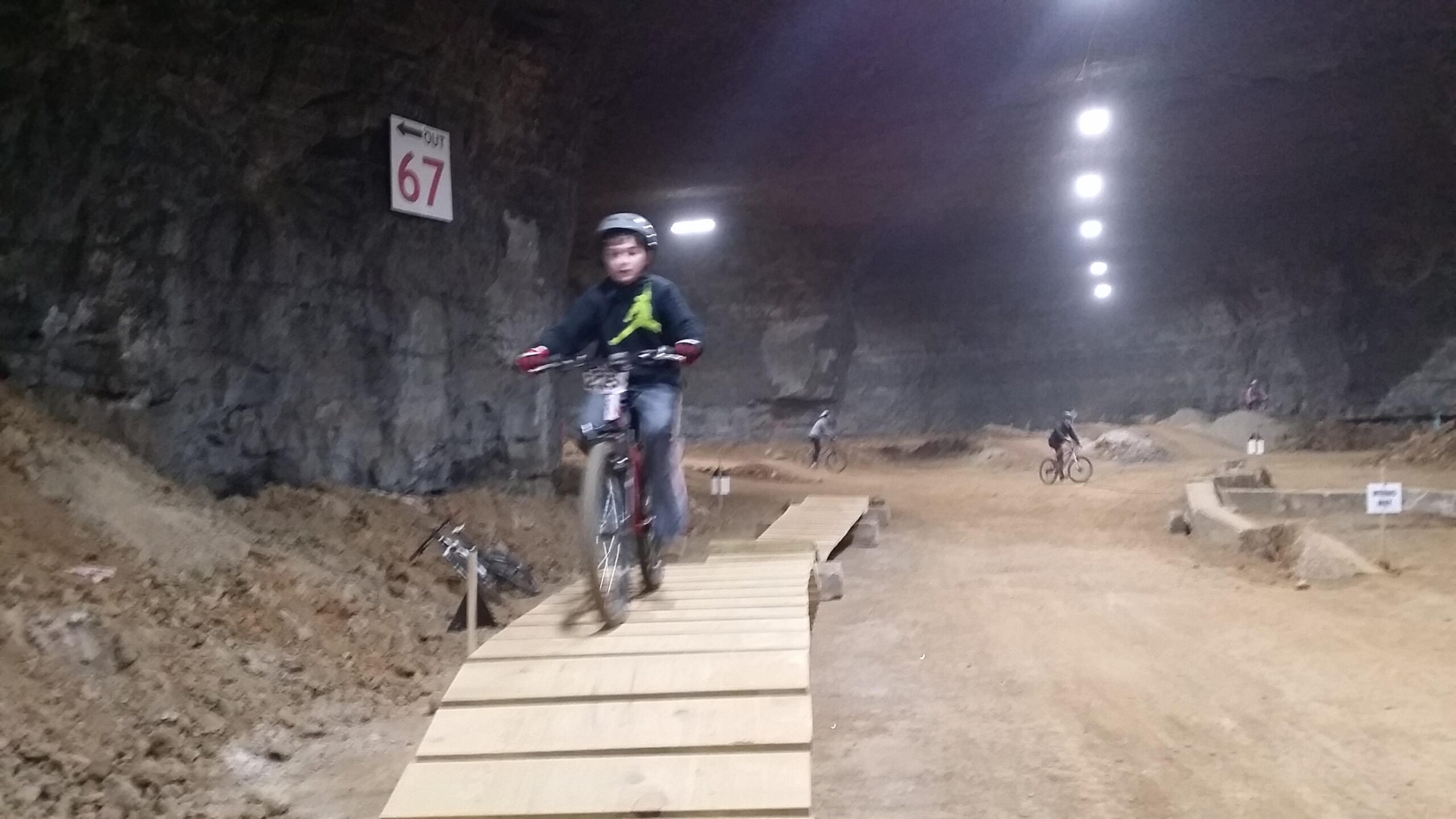 A young rider in a helmet navigates a wooden ramp on a mountain bike inside a large underground cavern, with rocky walls and artificial lighting. Other cyclists can be seen on the dirt track in the background. A sign marked "67" is visible on the wall, indicating an exit. Louisville Mega Cavern Bike Park mountain bike trail.