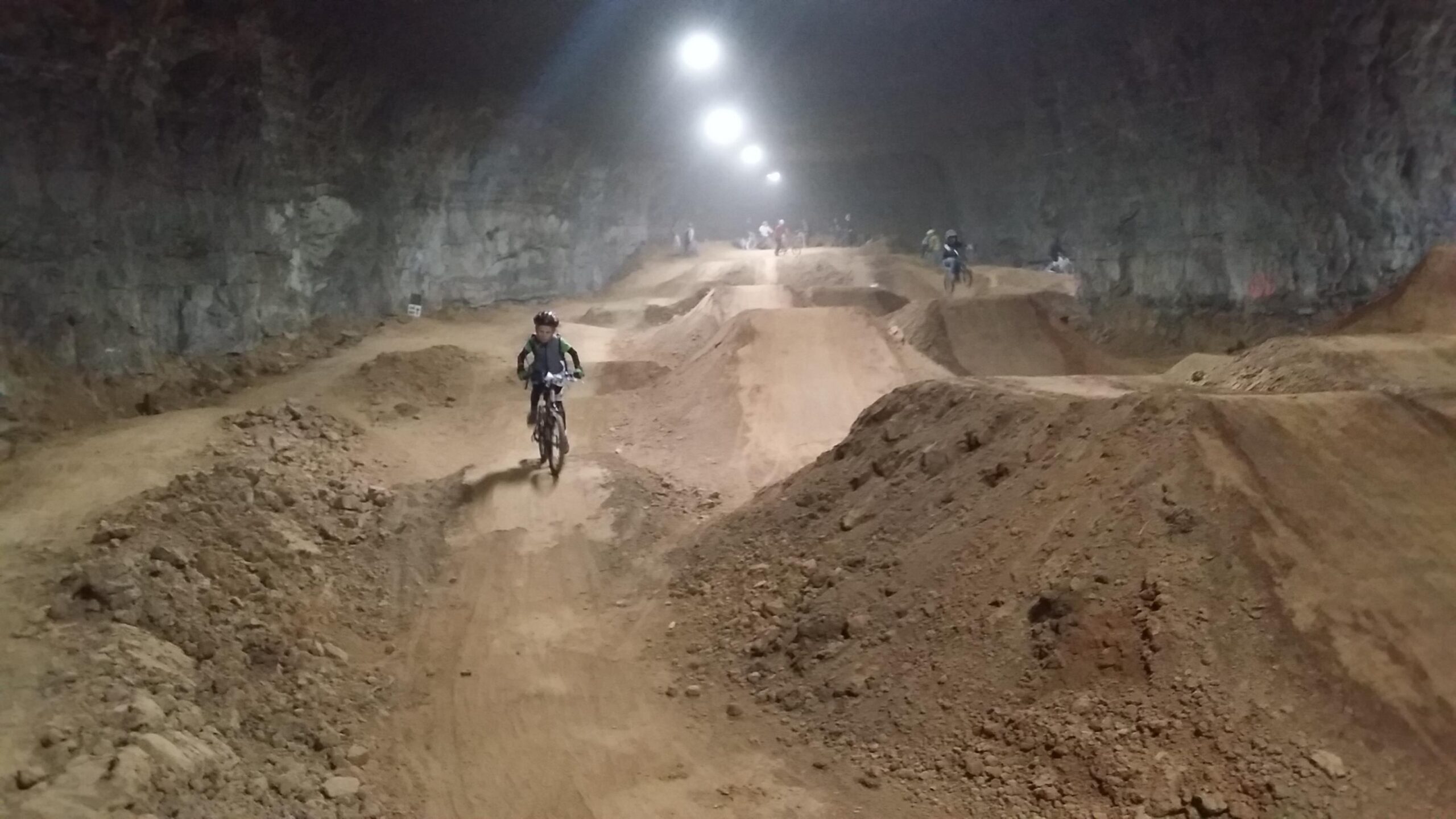 A cyclist rides a mountain bike on a dirt trail in a large underground cavern, with rocky walls and uneven terrain. The path features dirt mounds and jumps, and several other riders are visible in the background under bright overhead lights. Louisville Mega Cavern Bike Park mountain bike trail.