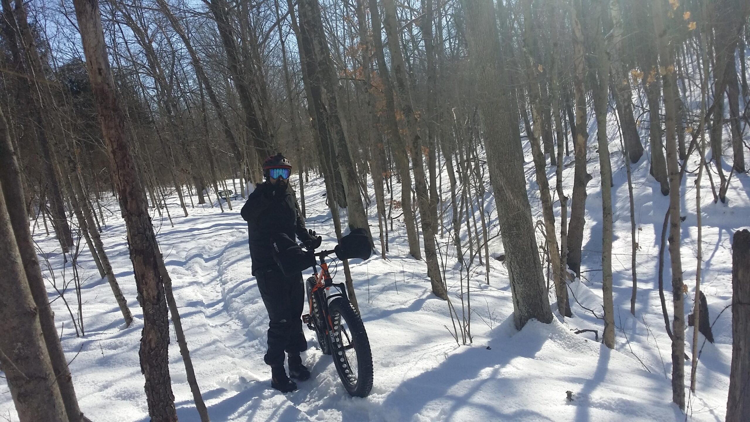 A person wearing a black winter outfit and goggles is standing next to a fat bike in a snowy forest. The snow covers the ground, and the trees are bare, indicating winter. Sunlight filters through the branches, creating a bright, clear day. England Idlewild Mountain Biking Park mountain bike trail.