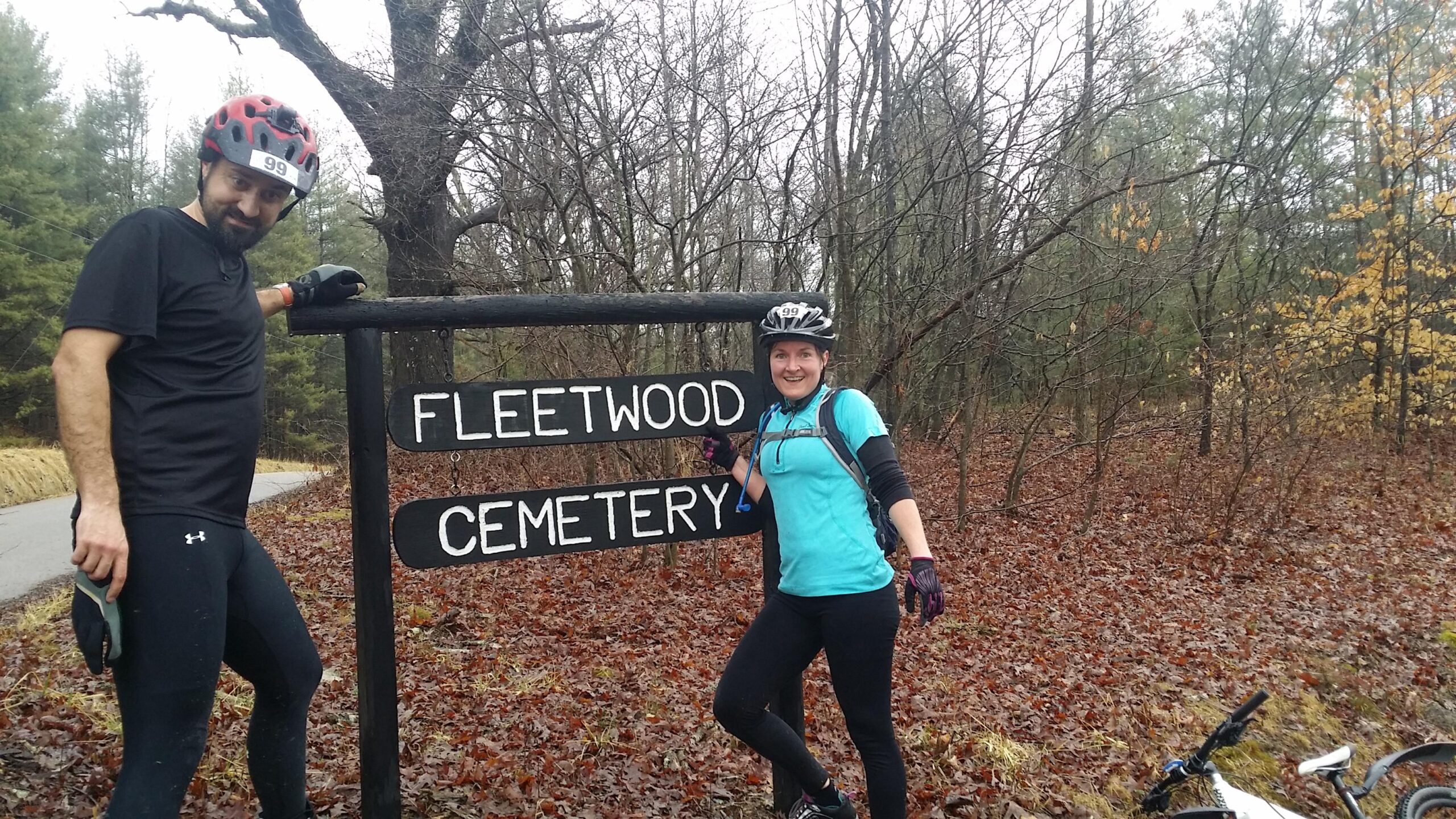 A man and a woman wearing cycling gear pose next to a wooden sign that reads "Fleetwood Cemetery." The background features a wooded area with bare trees and fallen leaves, suggesting a cloudy day. A bicycle is partially visible on the ground near them. Hickory Ridge mountain bike trail.