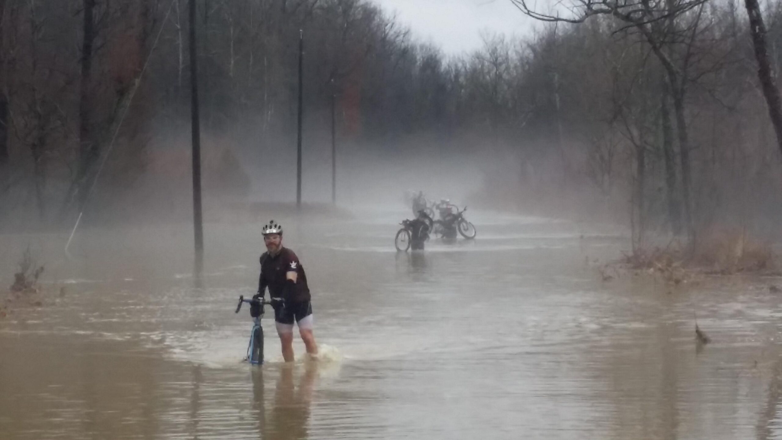 A man wades through a flooded street, pushing a bicycle, while several bikes are partially submerged in the water. The surrounding area is shrouded in mist, with bare trees lining the muddy path and electric poles visible in the background. Hickory Ridge mountain bike trail.