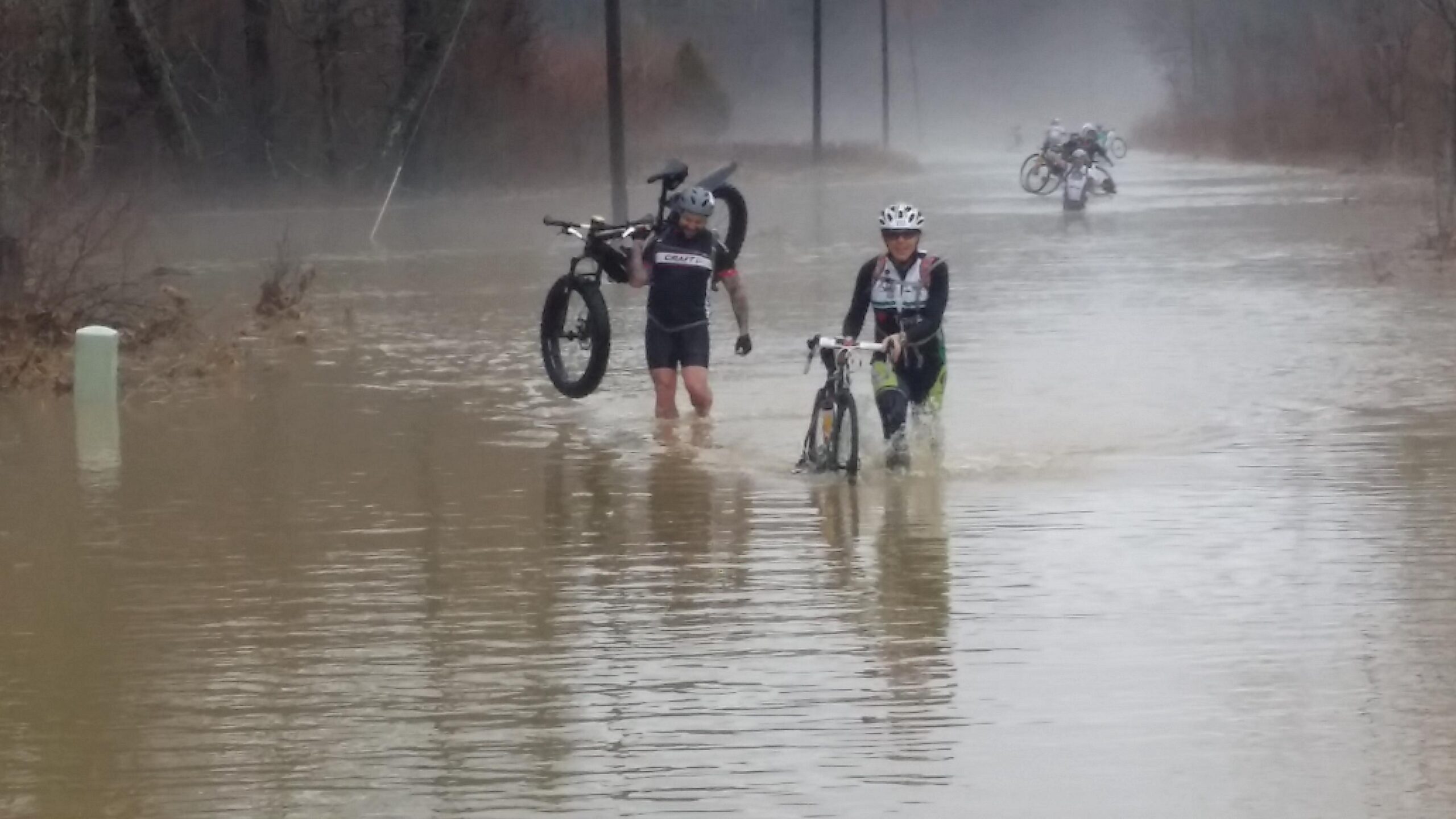 Two cyclists navigate a flooded area, each holding their bicycles above the water. The scene features murky water covering the ground, with trees and power lines visible in the background. The cyclists appear determined as they wade through the flood, showcasing the challenges of biking in adverse conditions. Hickory Ridge mountain bike trail.