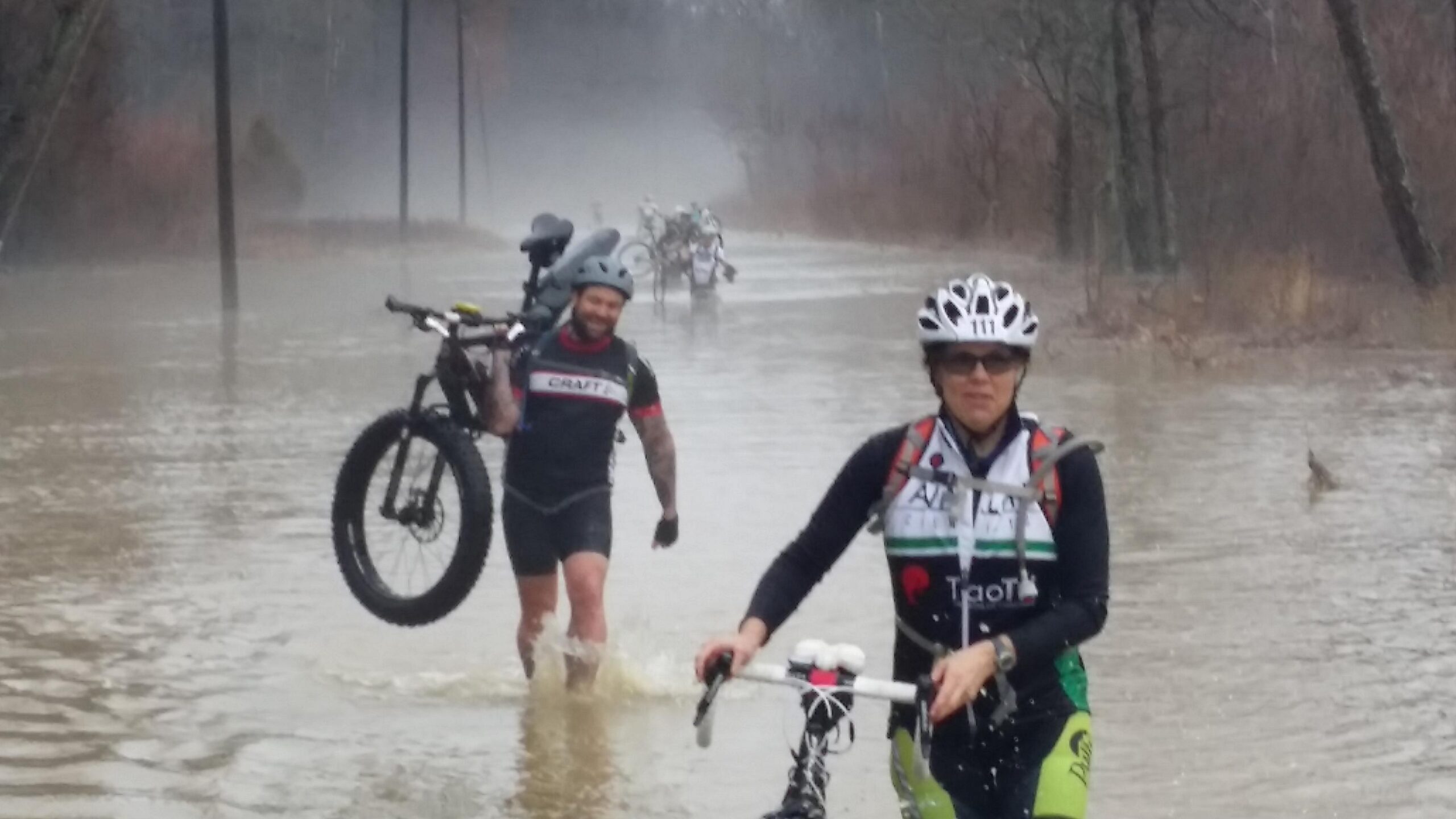 A group of cyclists wading through flooding, carrying their bikes in shallow water. The scene features a misty atmosphere with trees in the background and power lines visible. Two cyclists are prominently in the foreground, one smiling while carrying a bike and the other focused on navigating the water. Hickory Ridge mountain bike trail.