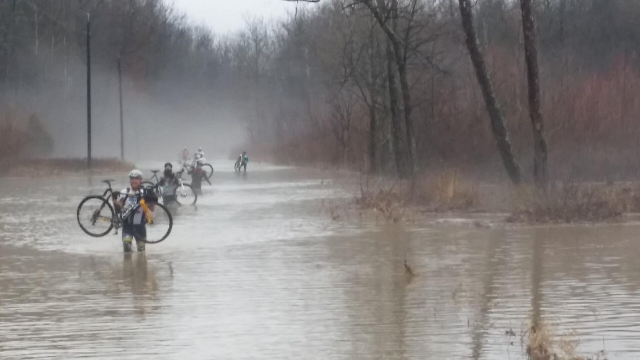 A group of cyclists wading through a flooded path, holding their bicycles above the water. The scene is misty with trees lining the background, and the atmosphere suggests recent rain. Hickory Ridge mountain bike trail.