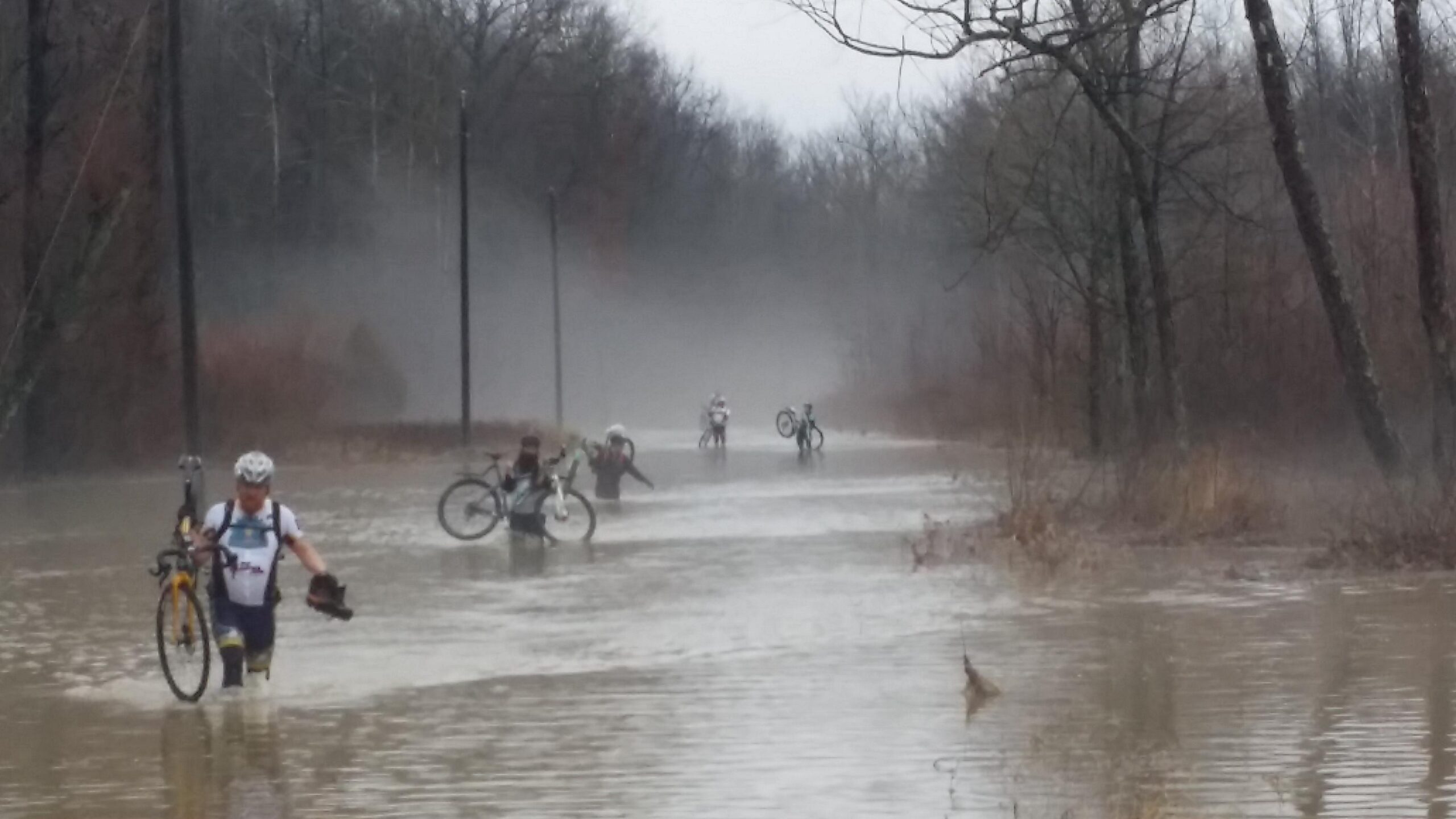 A group of cyclists wading through a flooded path, carrying their bikes as water reaches mid-calf. The surrounding area is blurry with mist and bare trees, indicating a rainy day. Hickory Ridge mountain bike trail.