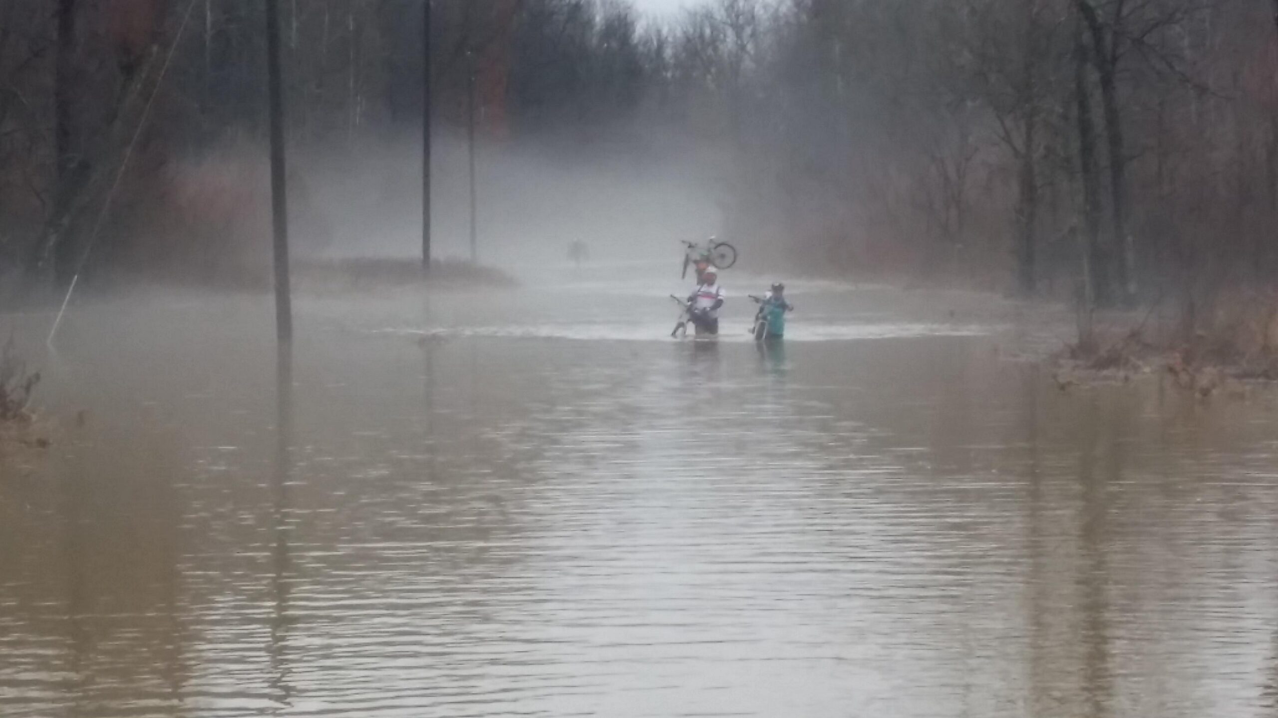 Two people wade through a flooded area, with water up to their waists. One person is holding a bicycle above their head. The background features fog and trees, creating a misty atmosphere, while power lines are visible along the edge of the water. Hickory Ridge mountain bike trail.