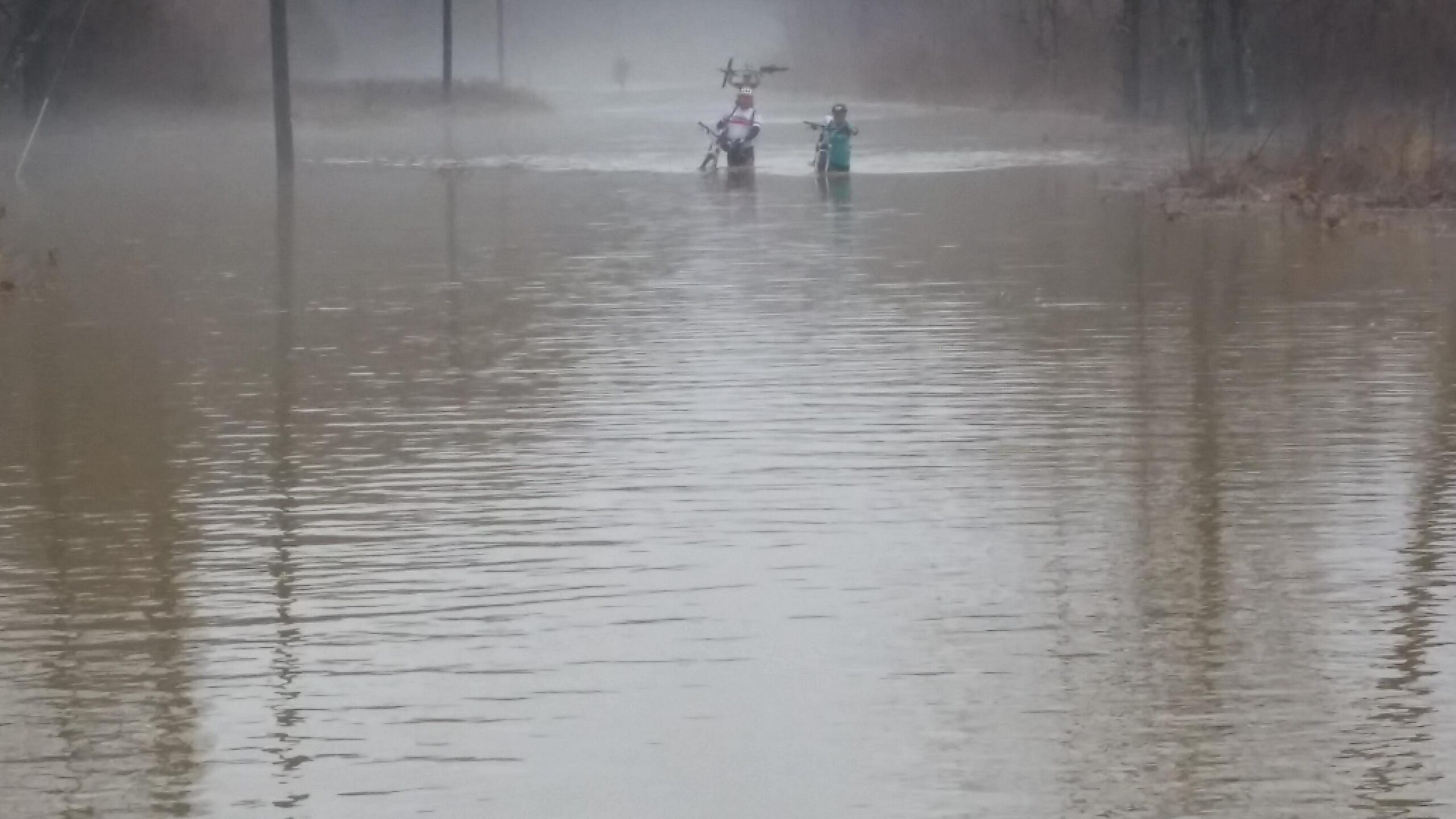 Two people wade through a flooded area on a misty day, surrounded by water that reflects the overcast sky. In the background, utility poles are partially submerged, indicating significant flooding in the region. The atmosphere is somber, with fog enveloping the landscape. Hickory Ridge mountain bike trail.